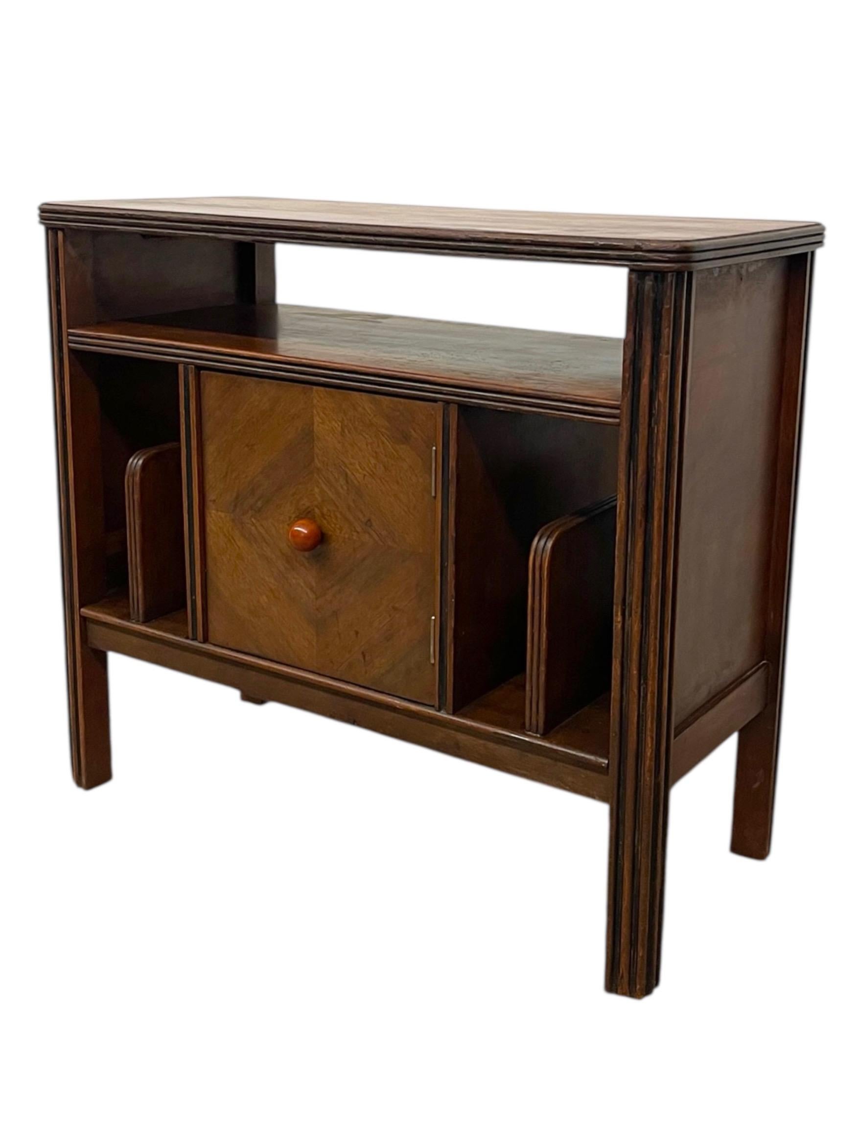 Un mobile Art Deco datato, realizzato con accenti lungo gli angoli anteriori scanalati. Il mobile presenta un piano rettangolare con bordi arrotondati, un ripiano aperto e un'anta centrale disposta a rombo, rifinita con un pomello rotondo.
Ai lati