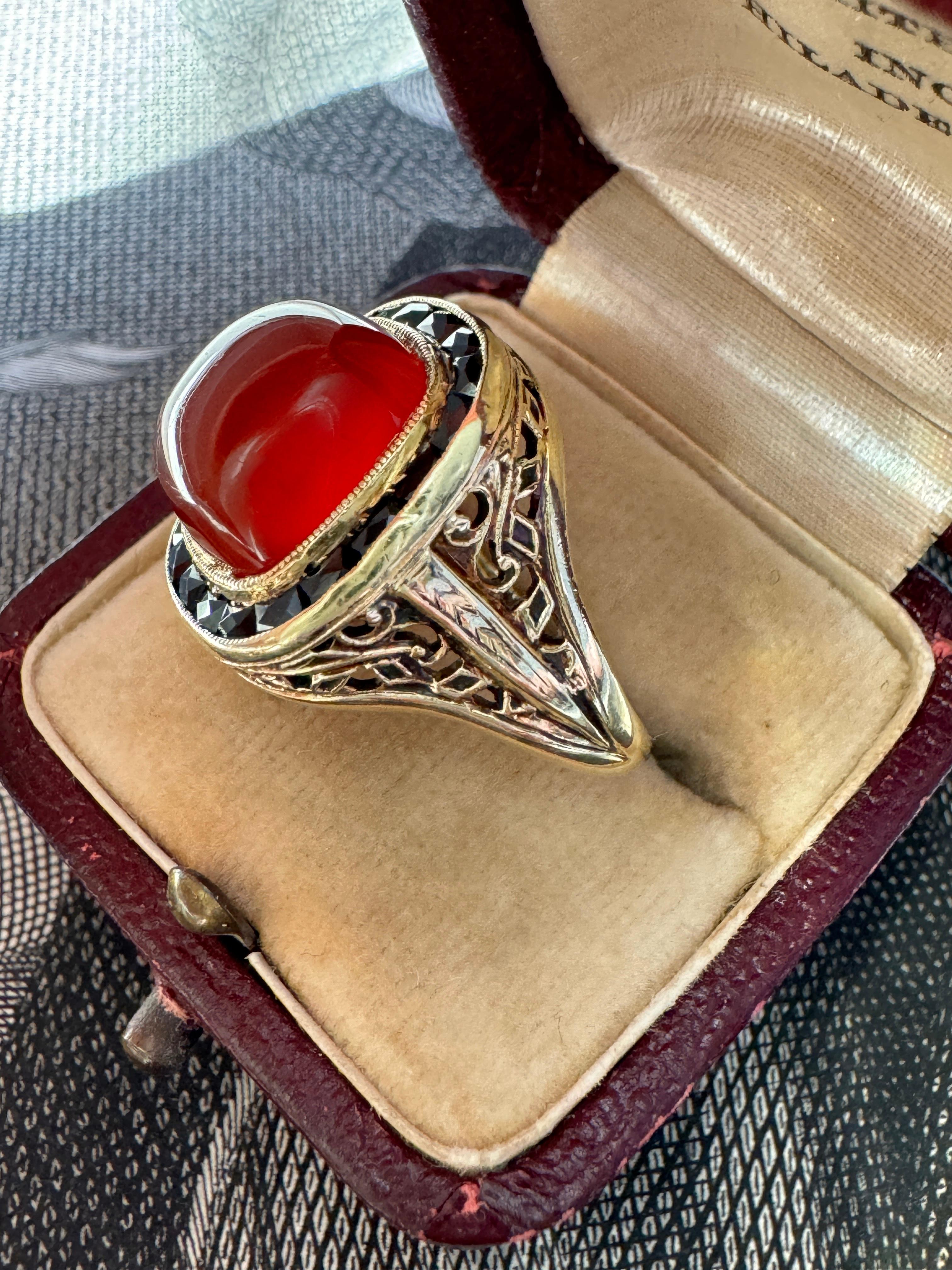 A glowing carnelian cabochon is dramatically framed by seamlessly set scissor-cut onyx in this 1930's ring by Bailey, Banks, and Biddle. Crafted in 14 karat gold, the sides are artfully embellished with pierced details and hand engraving. Currently