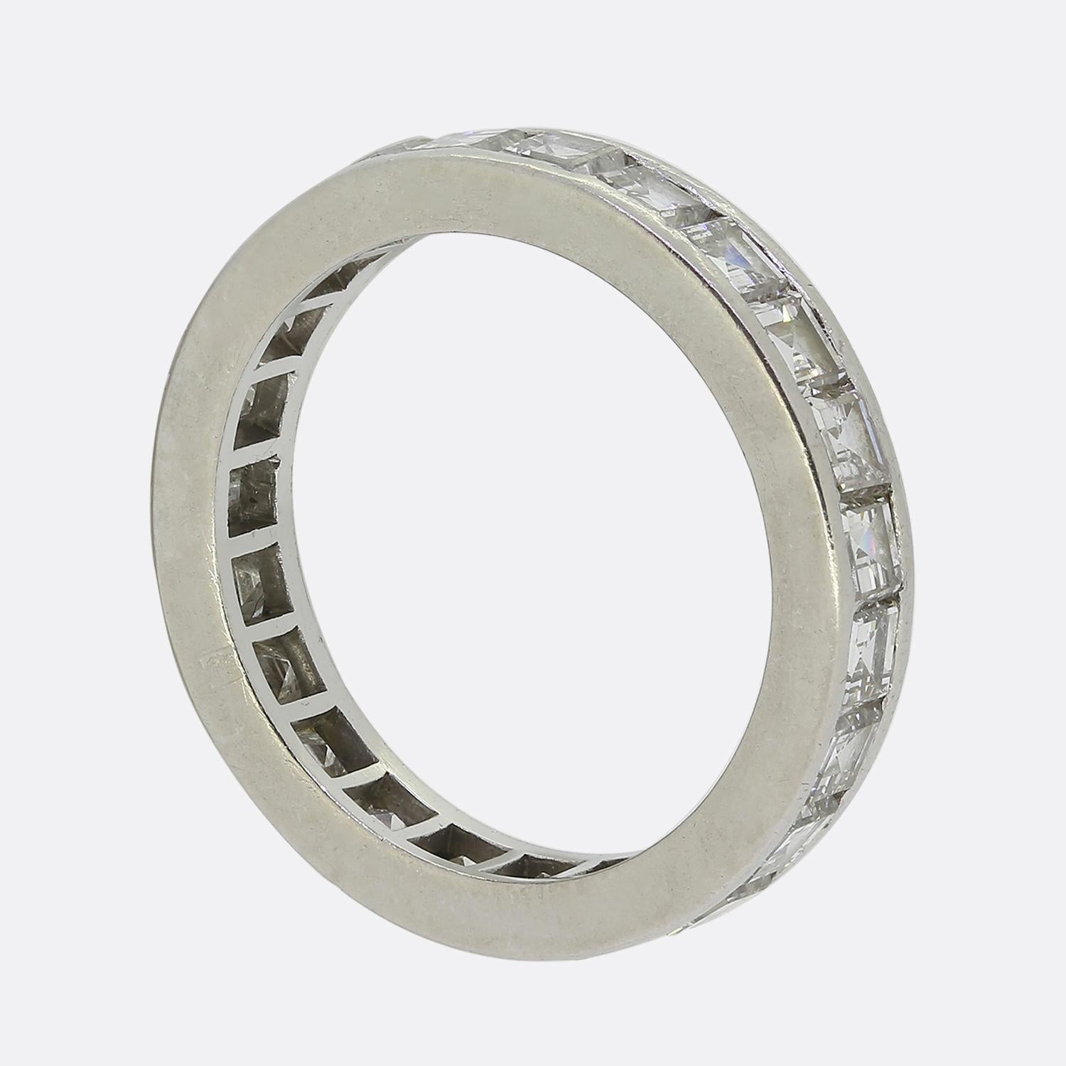 Here we have a stylish eternity ring crafted during the Art Deco movement. This platinum piece features a continuous row of carré-cut diamonds, each meticulously channel set within a sleek bordering above and below. The diamonds are arranged in a
