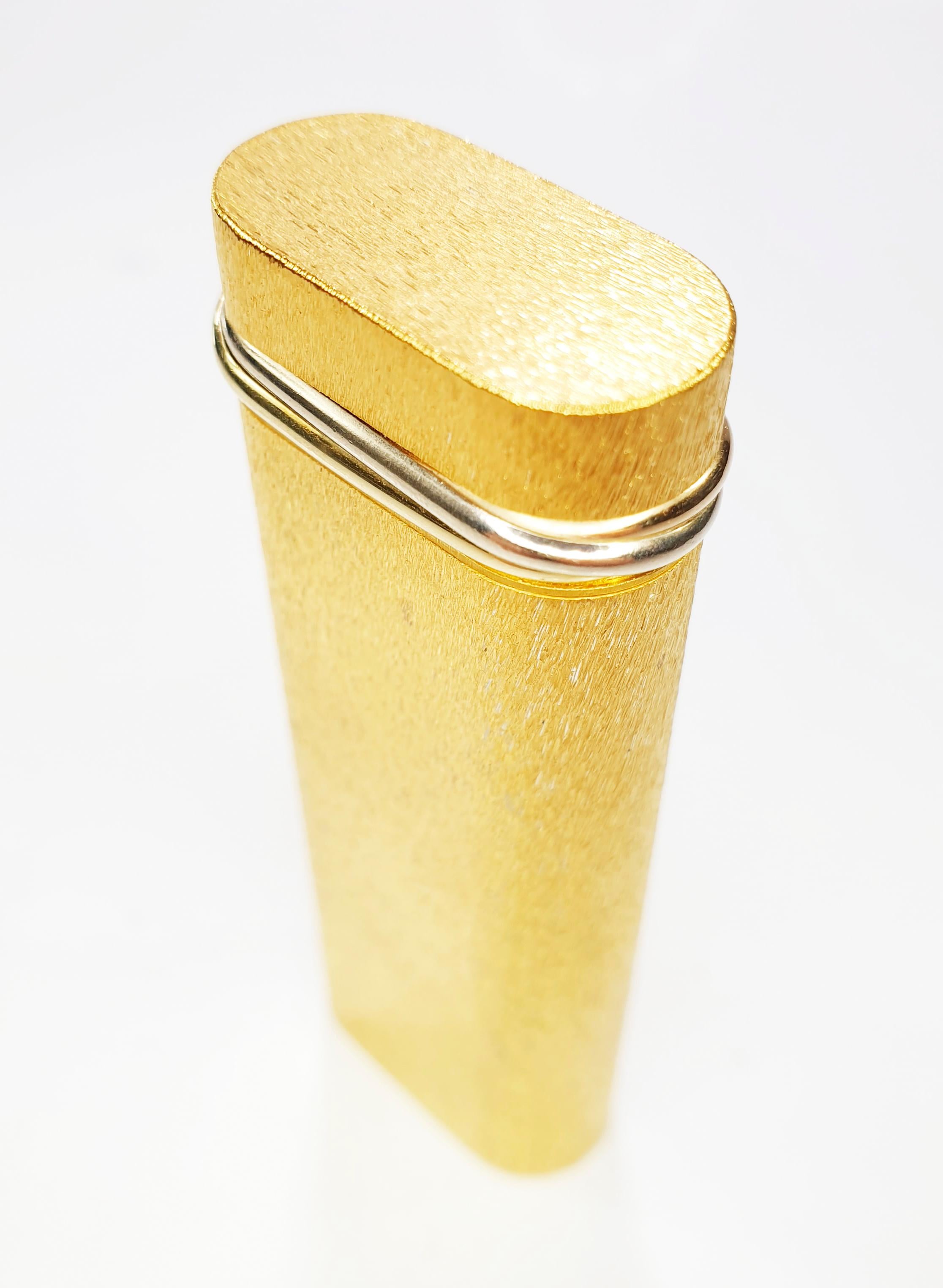 Pocket Trinity Cartier lighter  in 18-karat gold-plated textured finish with tri coloured gold collar an oval shaped body and a ribbed/fluted design. 
It has a flip-top lid that houses the Swiss-made mechanism. The flint wheel pivots when opened and