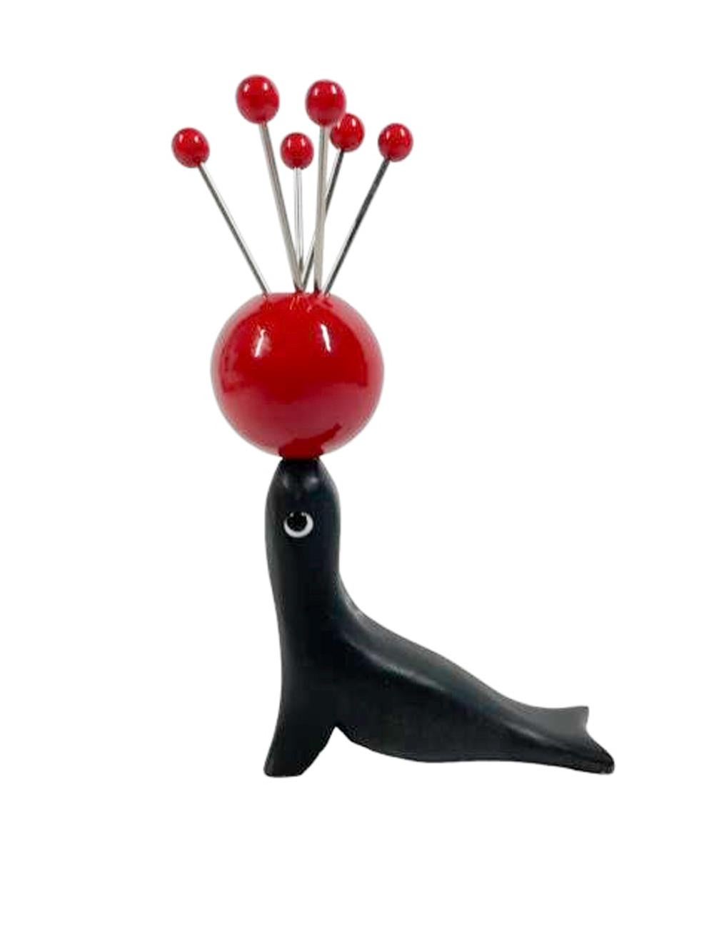 Juego de seis picos de cóctel con forma de bola sostenidos por una bola roja en equilibrio sobre la nariz de una foca con ojos de cristal. Púas de acero inoxidable con tapas de bolas de baquelita roja y soporte de madera tallada pintada.