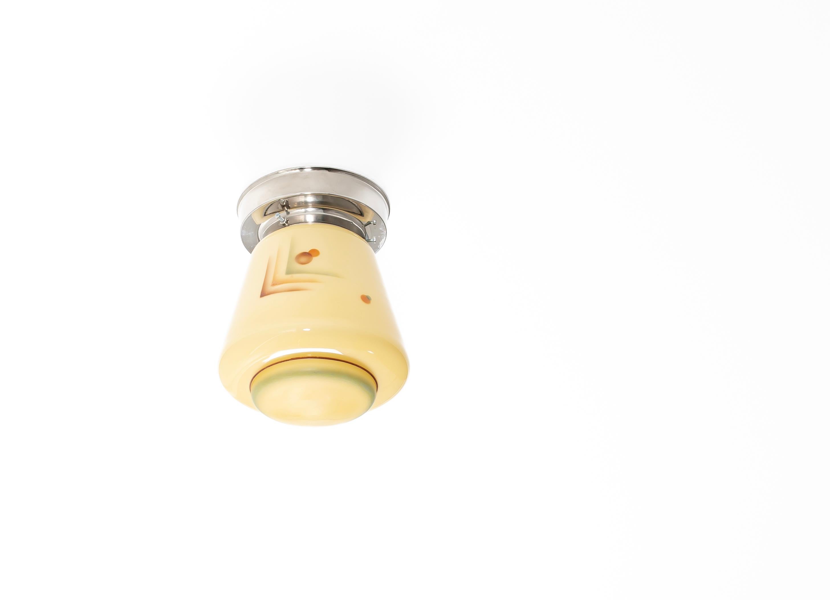 Bellissima e suggestiva plafoniera da incasso risalente agli anni '30, con un paralume in vetro dipinto a mano con ornamenti geometrici. La forma affusolata e cilindrica è realizzata in vetro opalino giallo crema e decorata con audaci motivi lineari