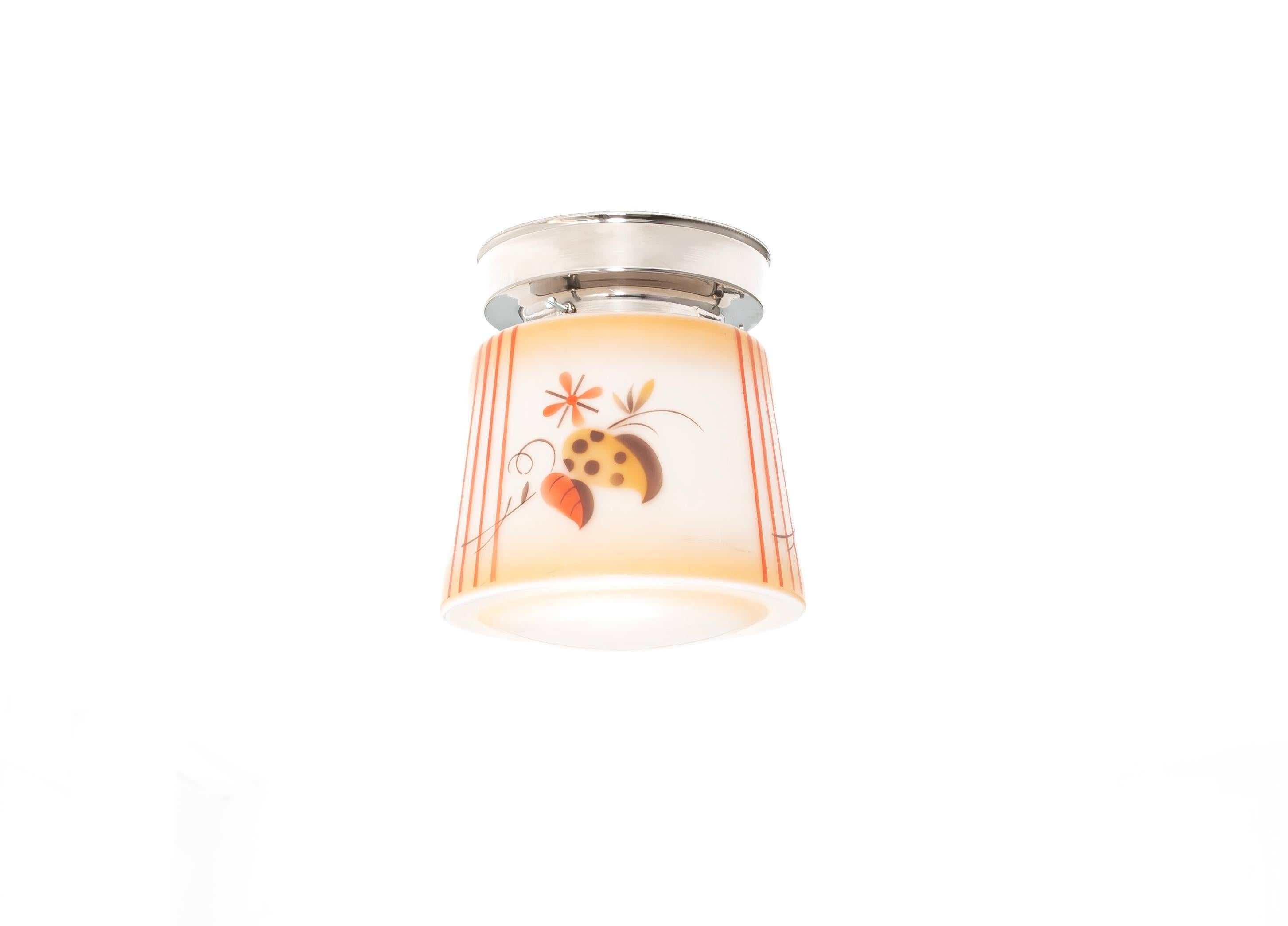 Maravillosa lámpara de techo empotrada que presenta una pantalla de cristal pintada a mano con un estilizado motivo floral y abstracto en cálidos tonos ámbar, naranja y marrón chocolate, delicadamente dispuestos sobre un suave fondo crema. Las rayas