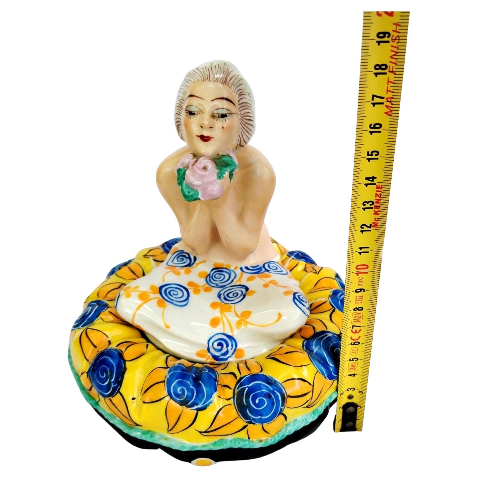 Poudrier en céramique Art Déco des années 1930, signé sous la base Aladin France - fabriqué en France. Whiting représente une jeune femme aux couleurs vives avec sa coupe de cheveux bob blanc et tenant un bouquet de fleurs, assise sur un coussin