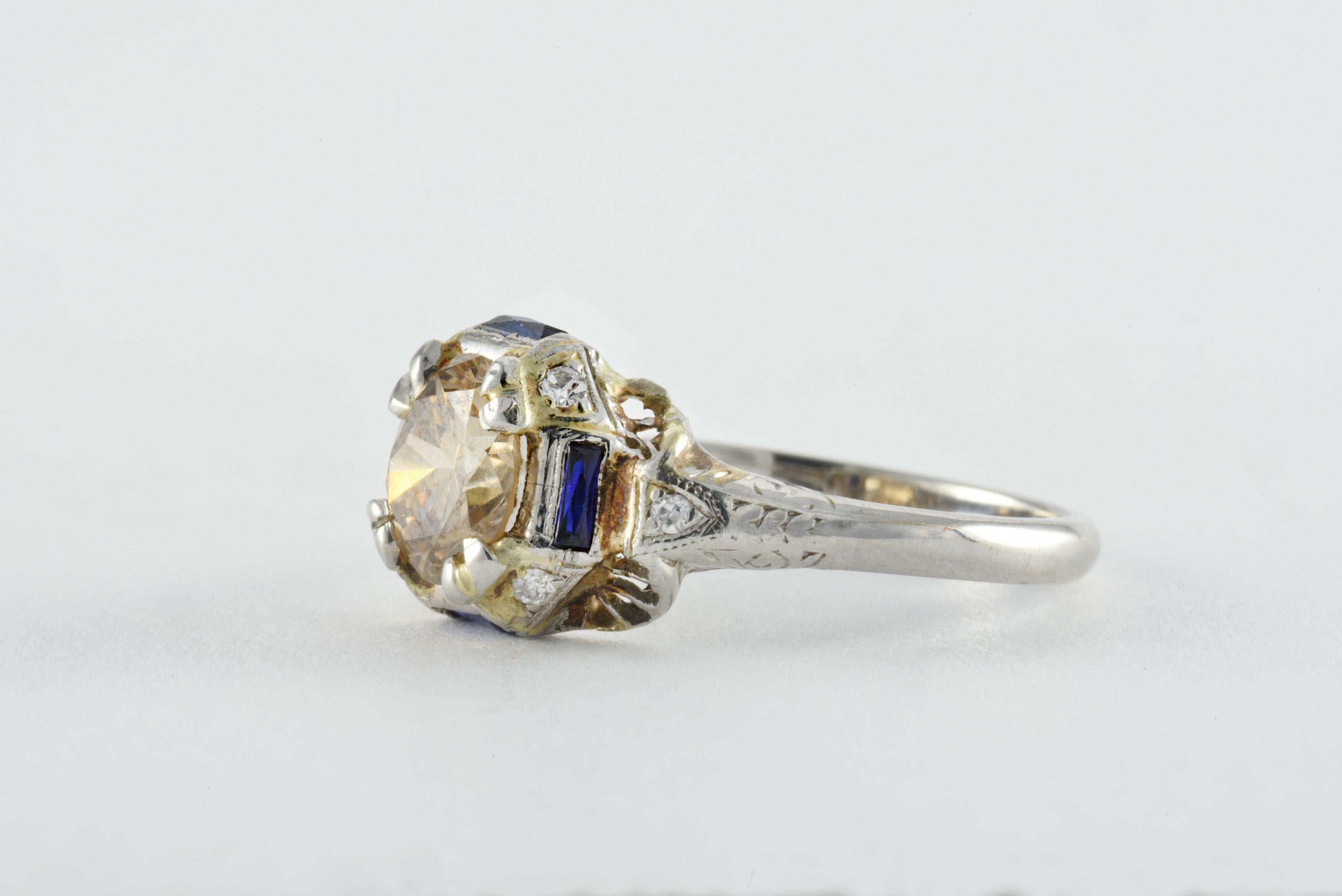 Crafted in the 1920s from 14kt white gold, this Art Deco band is designed around a bold Old European cut champagne colored diamond measuring approximately 1.34 carats accented by four baguette-cut blue sapphires, four single cut diamonds, and