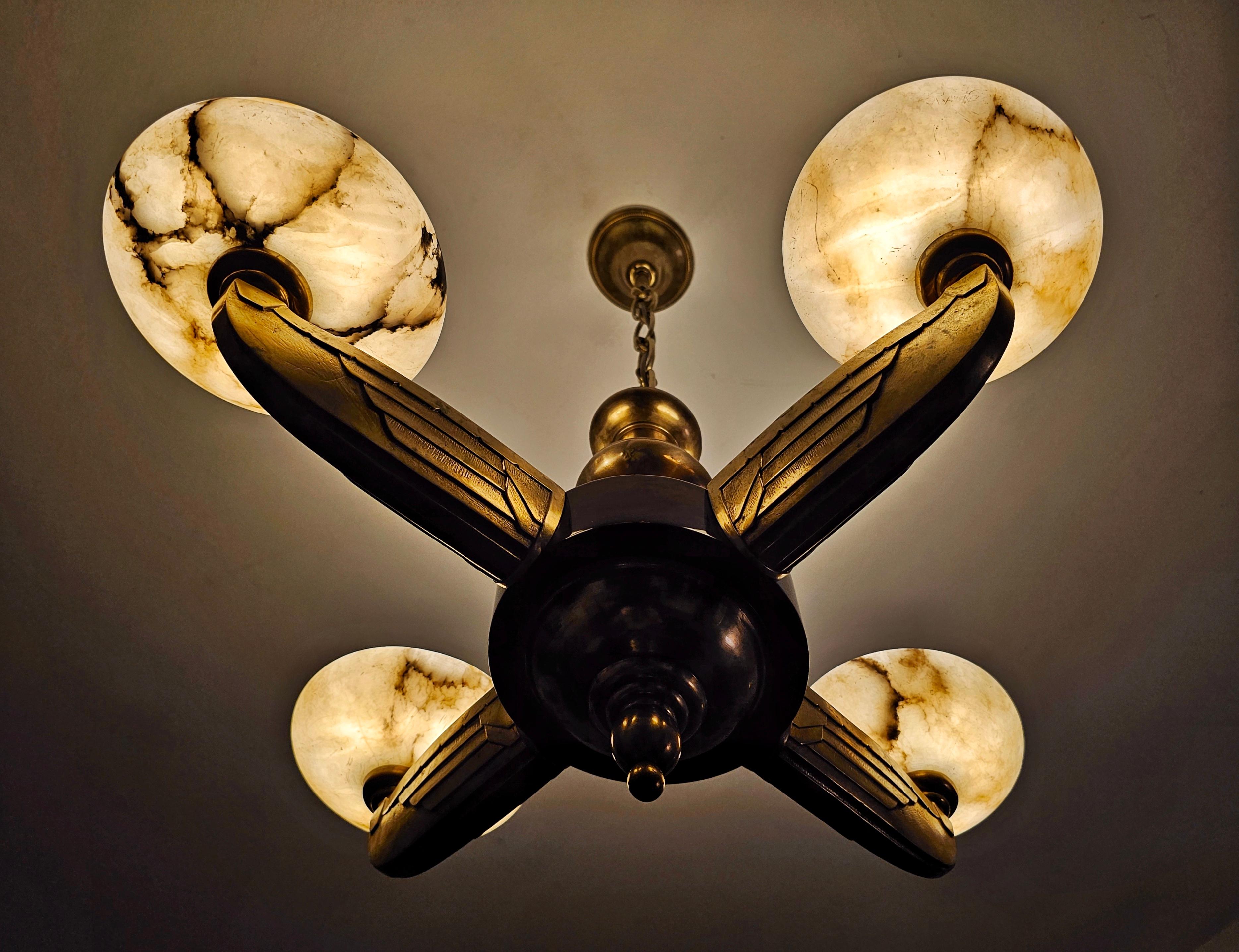 Dans cette annonce, vous trouverez un lustre Art Déco exceptionnellement rare et de grande taille à 4 bras de lumière. Les bras du lustre sont en bronze moulé, attachés à une base en noyer, tandis que les abat-jours sont en albâtre. La conception et
