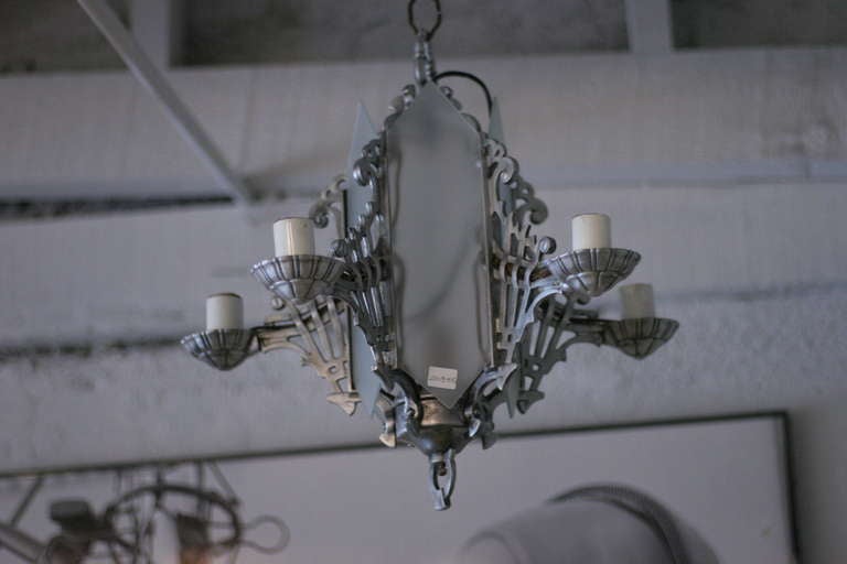 The five arms of this chandelier extend from the base of a glass paneled pendant. The piece features scrolling details throughout while the base of the chandelier features a geometric pattern.