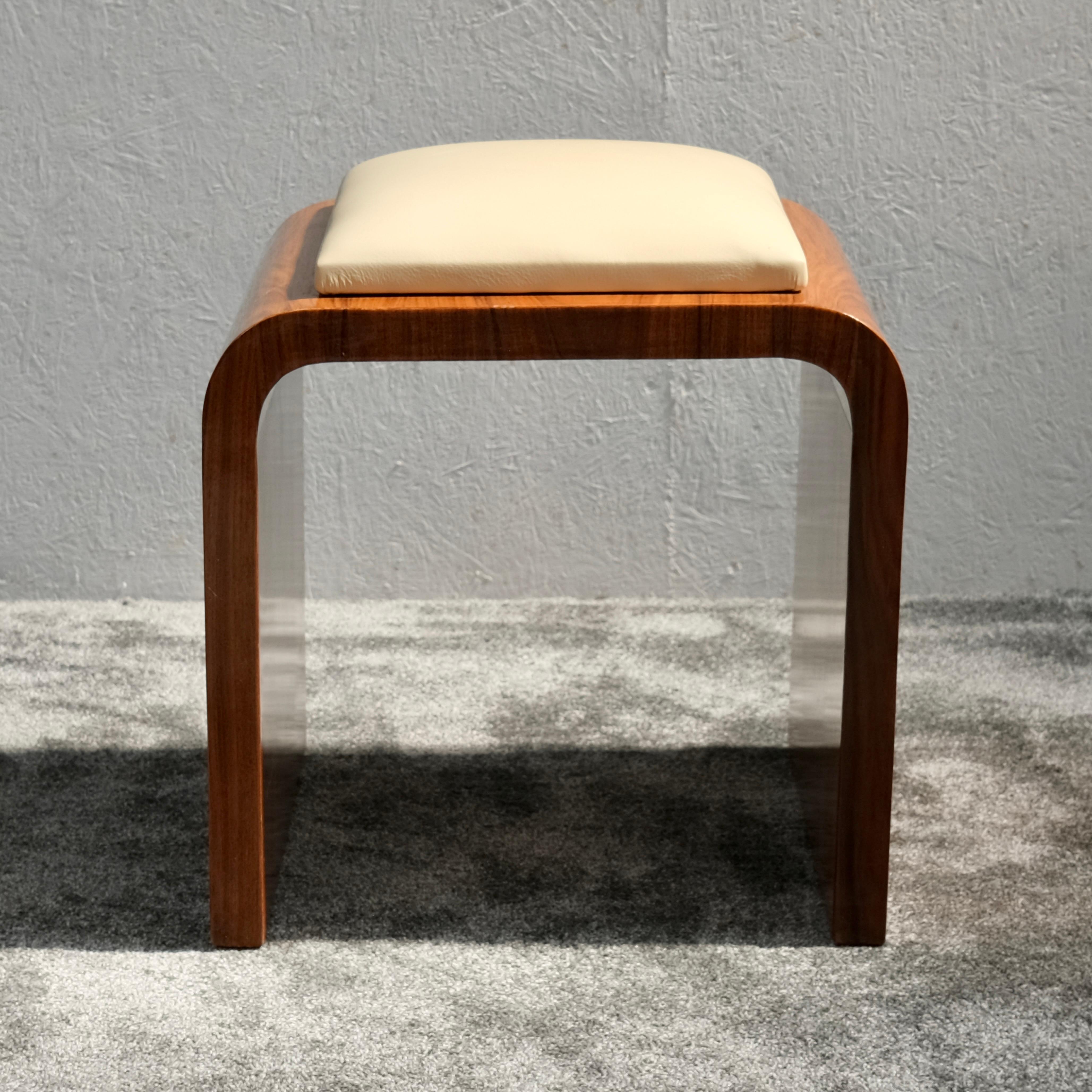 Ein charaktervoller Art-Déco-Pouf aus den 1930er Jahren, gefertigt aus reichem Kirschholz mit weichem, satiniertem Glanz und eleganter Maserung. Seine sanft geschwungene Silhouette wird von einem hellbeigen Lederkissen gekrönt, das dezente