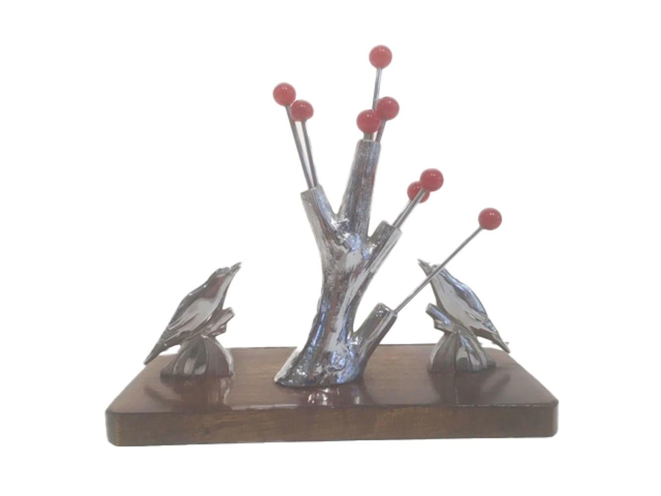 Eight red ball-topped chrome cocktail picks in a tree trunk stand where the pick-tops appear to be berries - two birds are at the base of the tree looking up at the berries. The chrome tree and birds are attached to a wooden base.