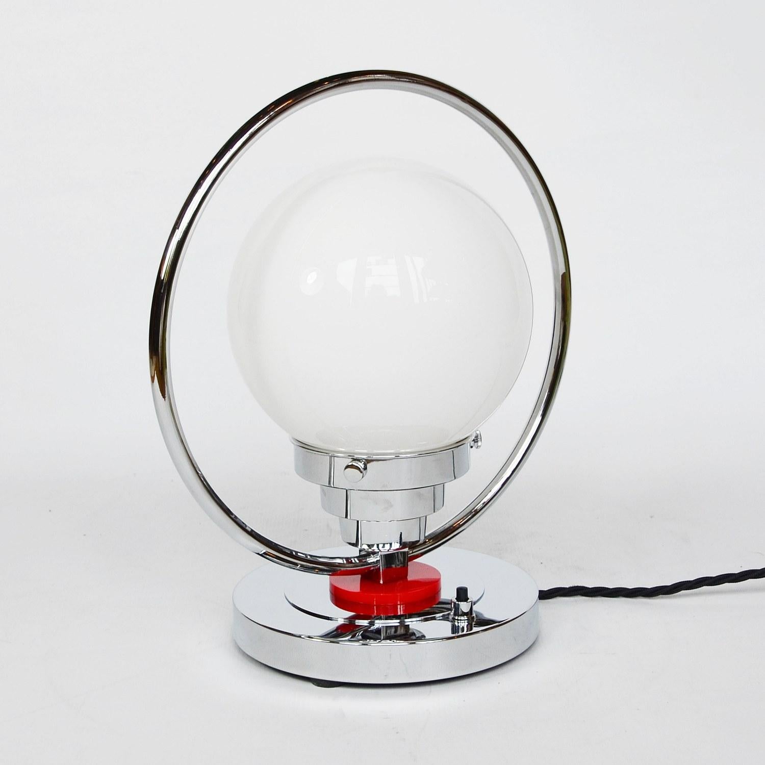 An Art Deco table lamp. Chromed metal base with red Catalin disc and mounted chromed hoop enclosing white glass shade.

Dimensions: H 20cm, W of shade 25cm, W of base 15cm

Origin: English

Item No: 210202

All of our lighting is fully