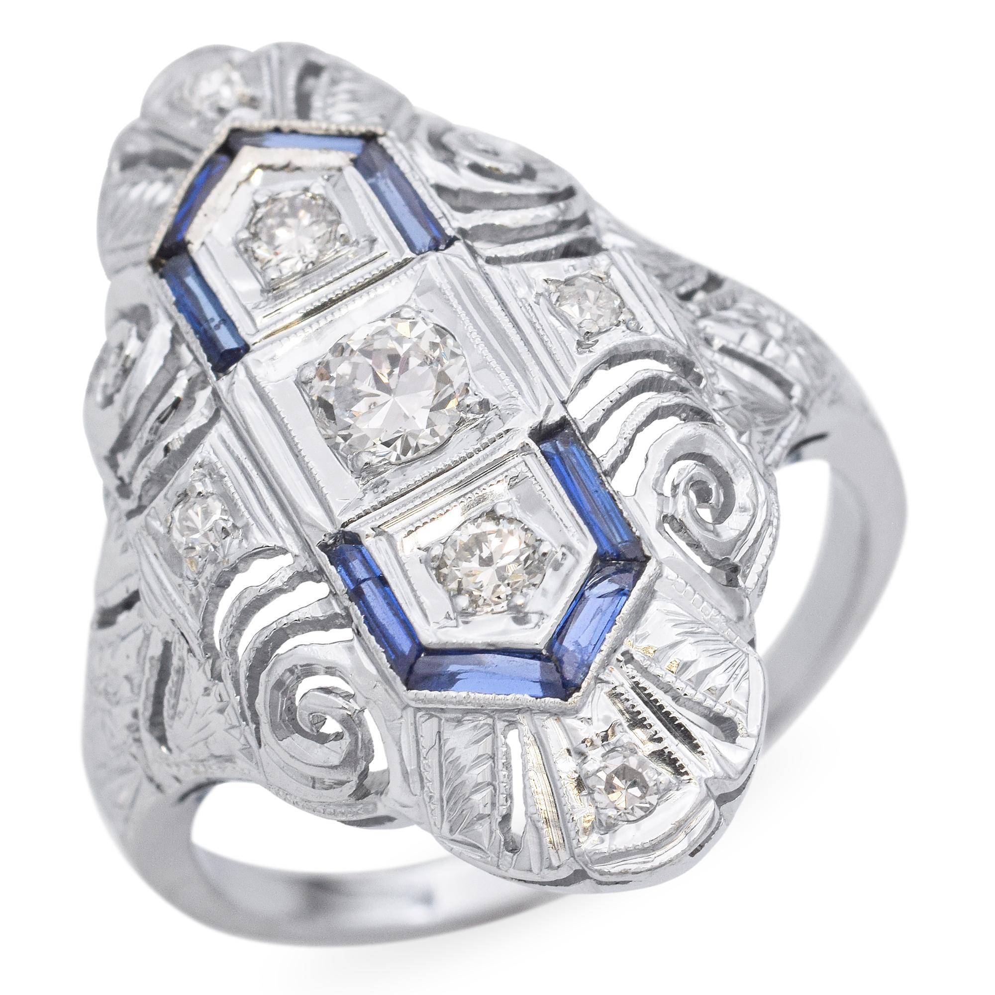 The ring features three central diamonds, each set within a geometric frame. Surrounding the diamonds are baguette-cut blue sapphires, adding a striking contrast. The frame is adorned with detailed scrollwork and additional small