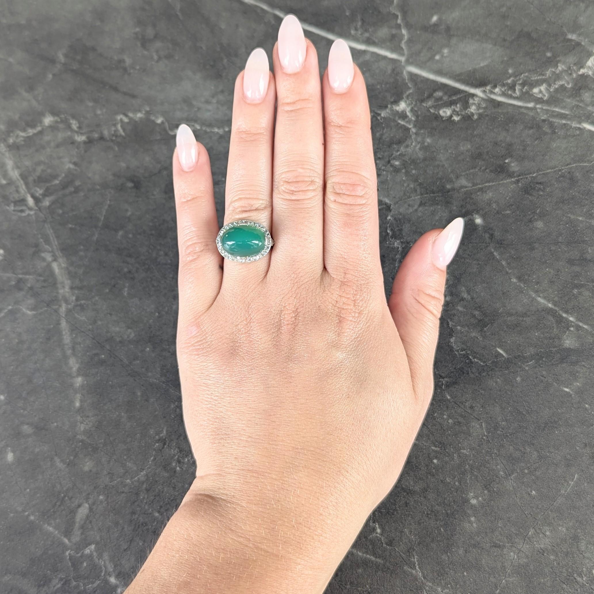 Centering an oval shaped chrysoprase cabochon measuring 10.0 x 15.0 mm
Translucent vibrant medium bluish green in color and set with tab prongs
With a recessed bead set halo surround of rose cut diamonds
With additional bead set diamonds on