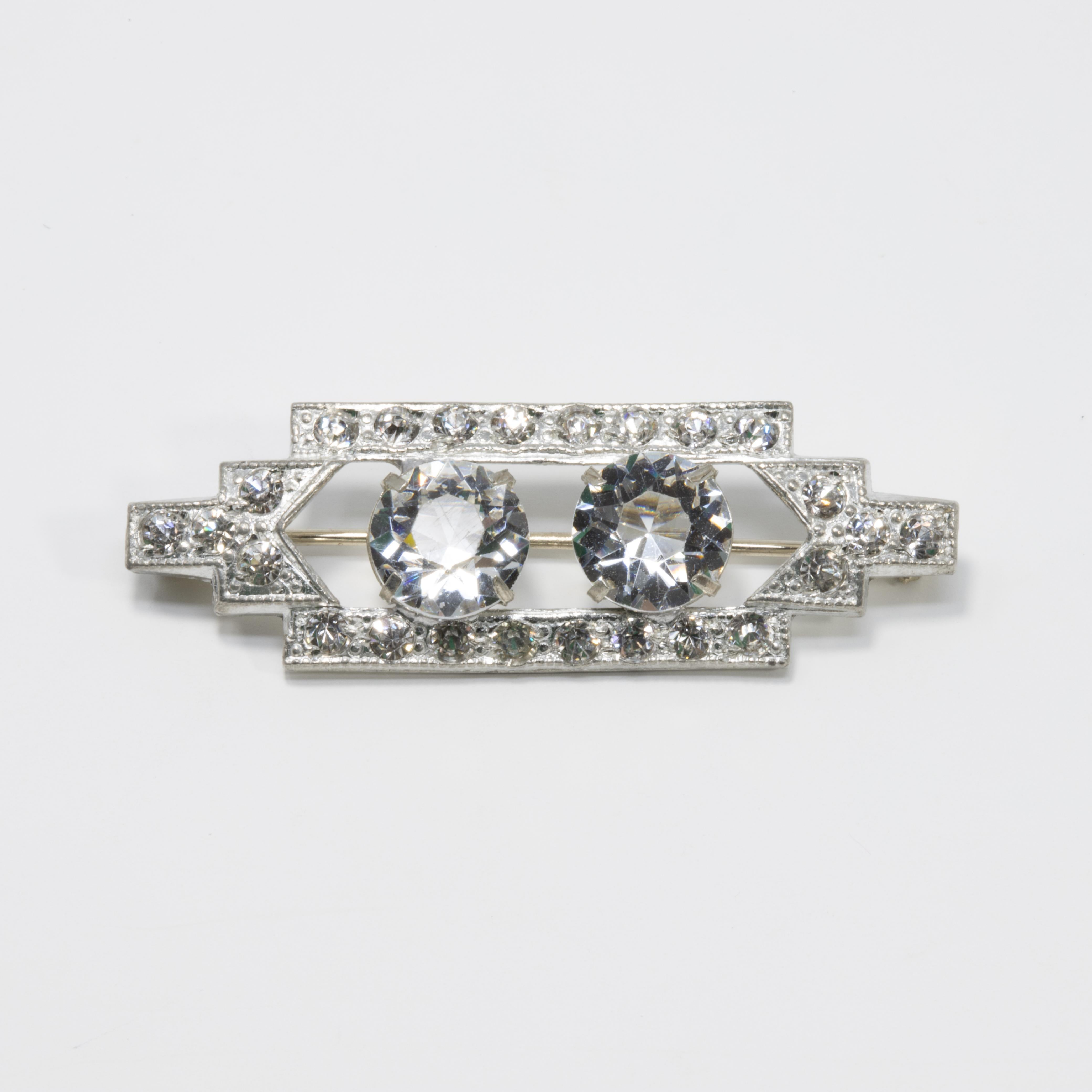 A sparkling art-deco pin from the early 1900s. This brooch features two larger clear crystals prong set in the center, surrounded by a rectangular silvertone setting accented with small crystals.