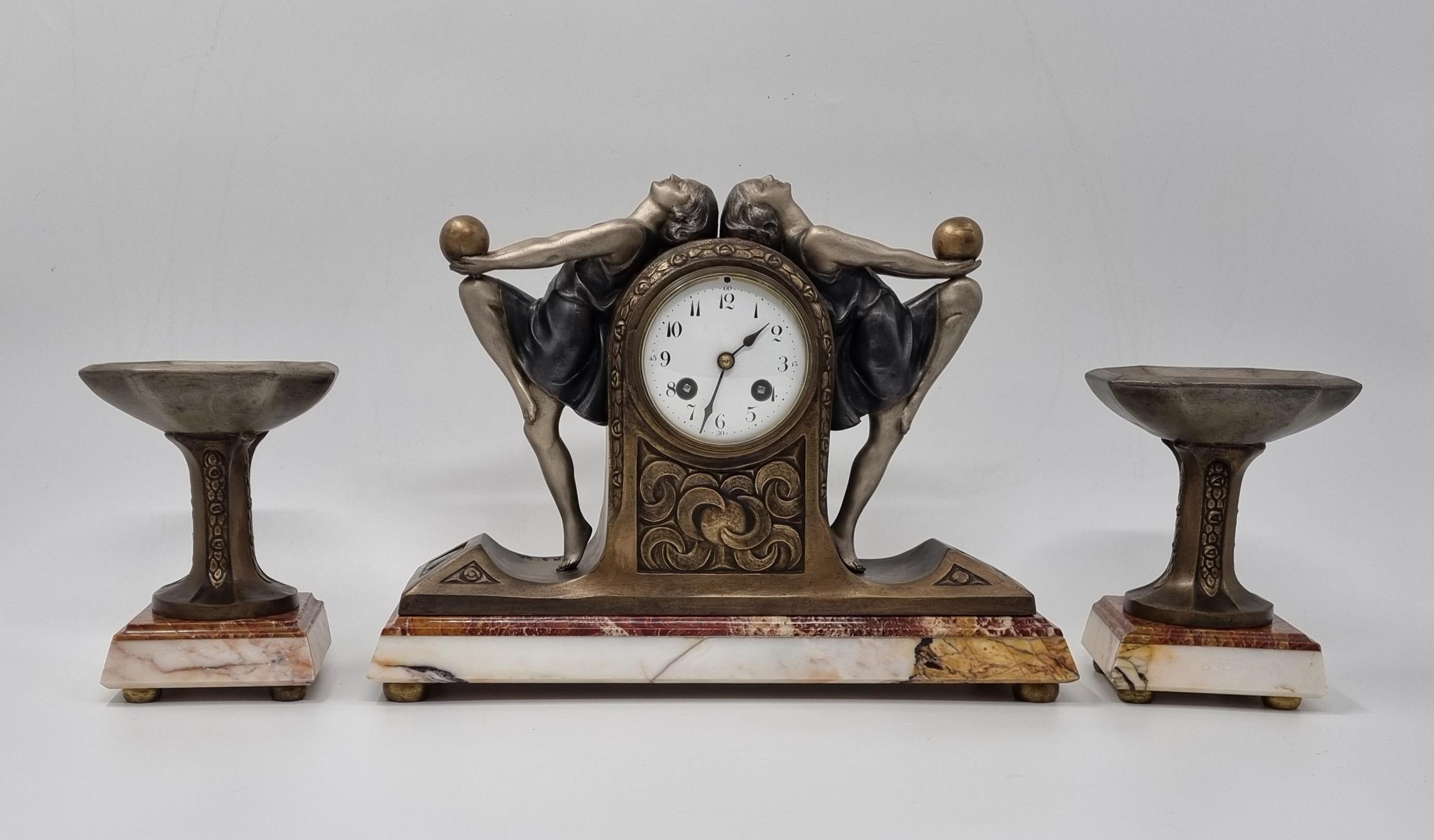 An Art Deco figural clock garniture set, circa 1920s-30s, Two daring Art Deco ball dancers elegantly positioned either side of the white enamel dial. Casy in cold painted spelter, mounted of coloured marble base. 

Signed P. Sega

In good, vintage