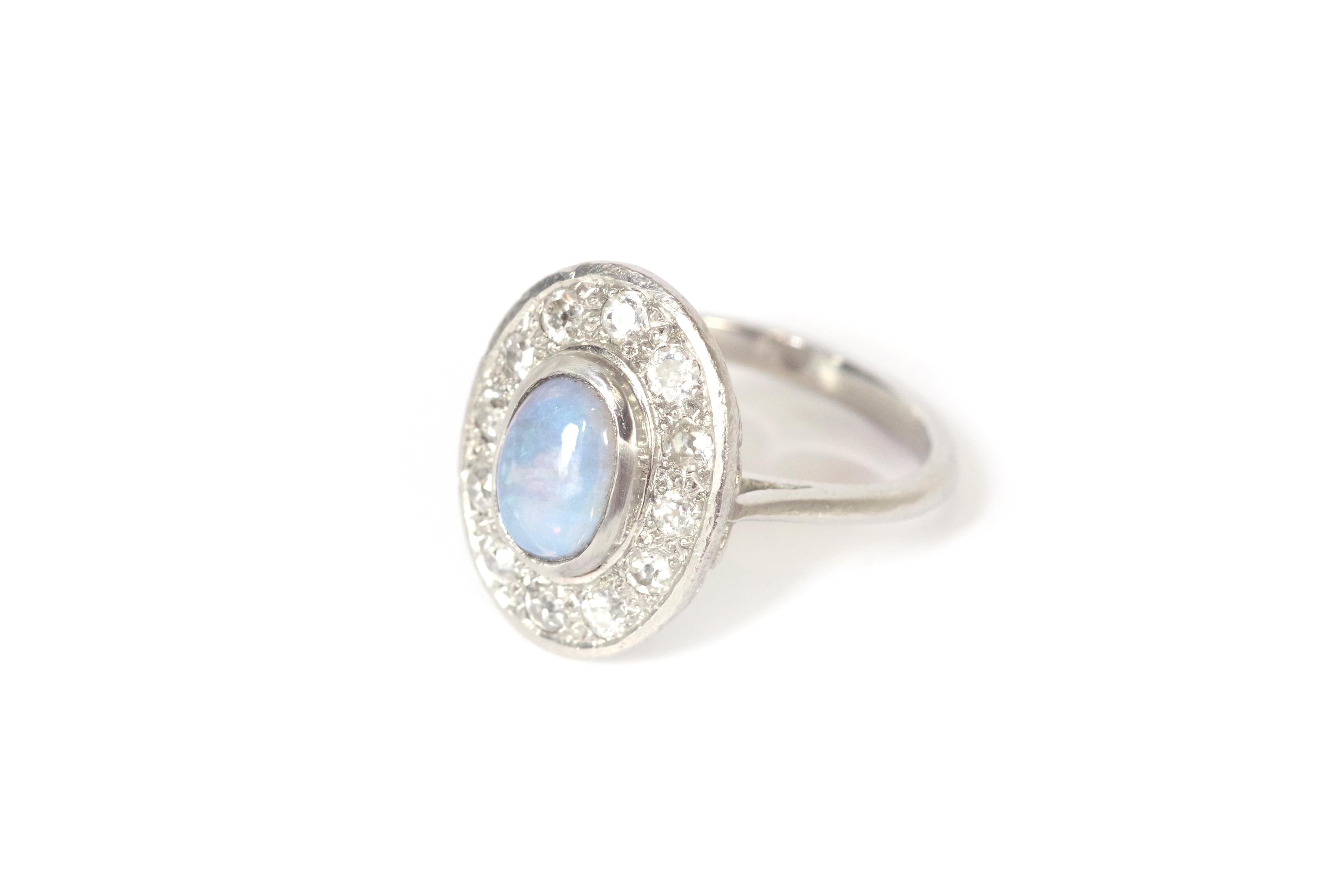 Art Deco cluster opal ring in platinum. This antique cluster ring features a central cabochon-cut opal set in a close setting, surrounded by twelve old-cut diamonds. The opal shows vibrant play-of-color with shades of violet, blue, and green,