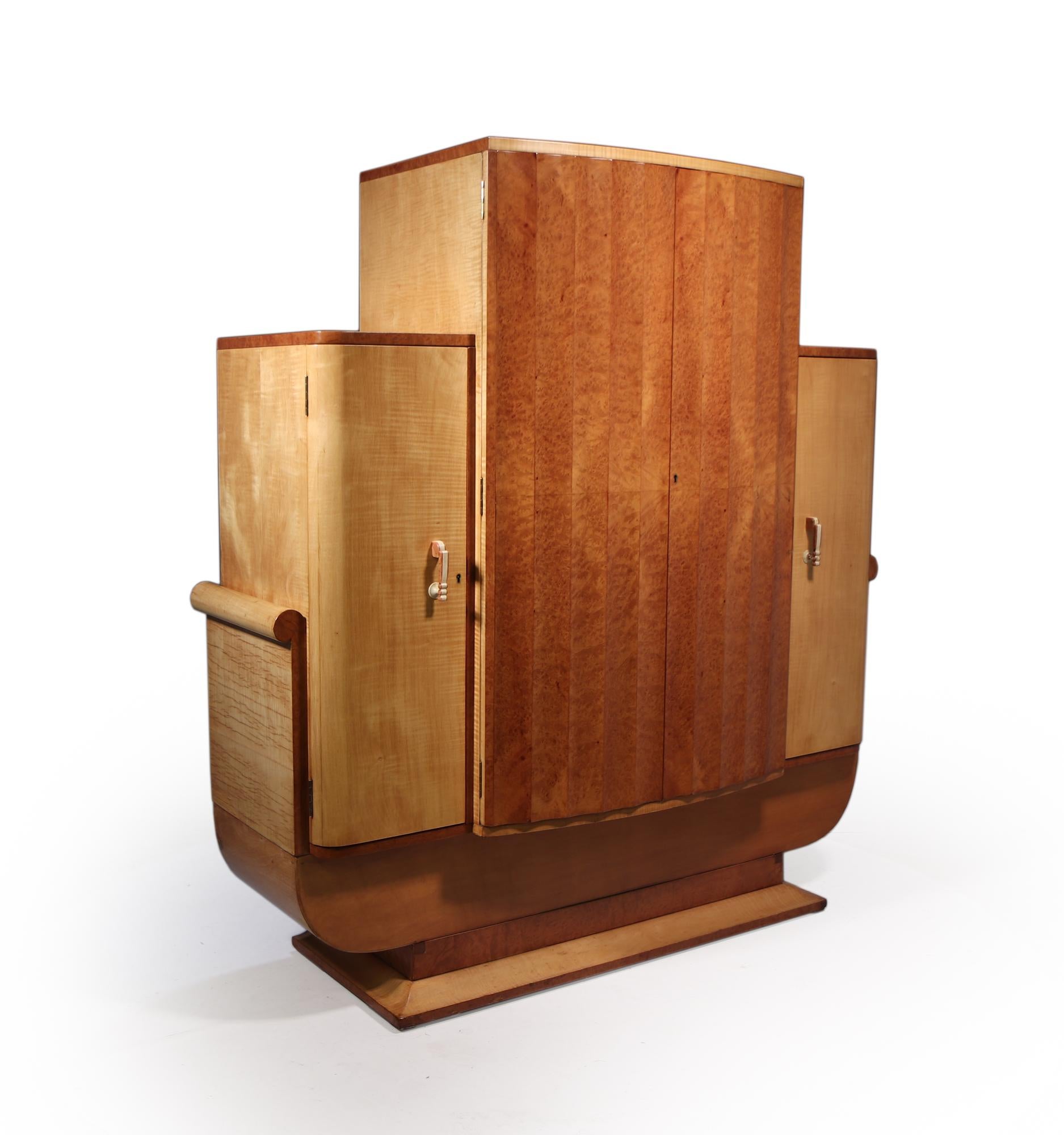 A design masterpiece designed and produced by by Harry and Lou Epstein in London in the 1930’s, this example in sycamore and burr walnut having fluted center doors that open to reveal cocktail making area with mirrored back and slide below is