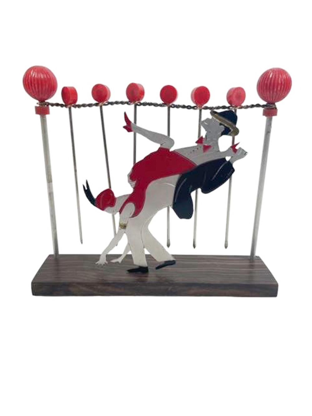 Plettri da cocktail e supporto in stile Art Deco. Una silhouette cromata dipinta di una coppia che fa Charleston davanti a una coppia di pali a sfera che sostengono un filo intrecciato che regge sei cocktail picks su una base di legno. 