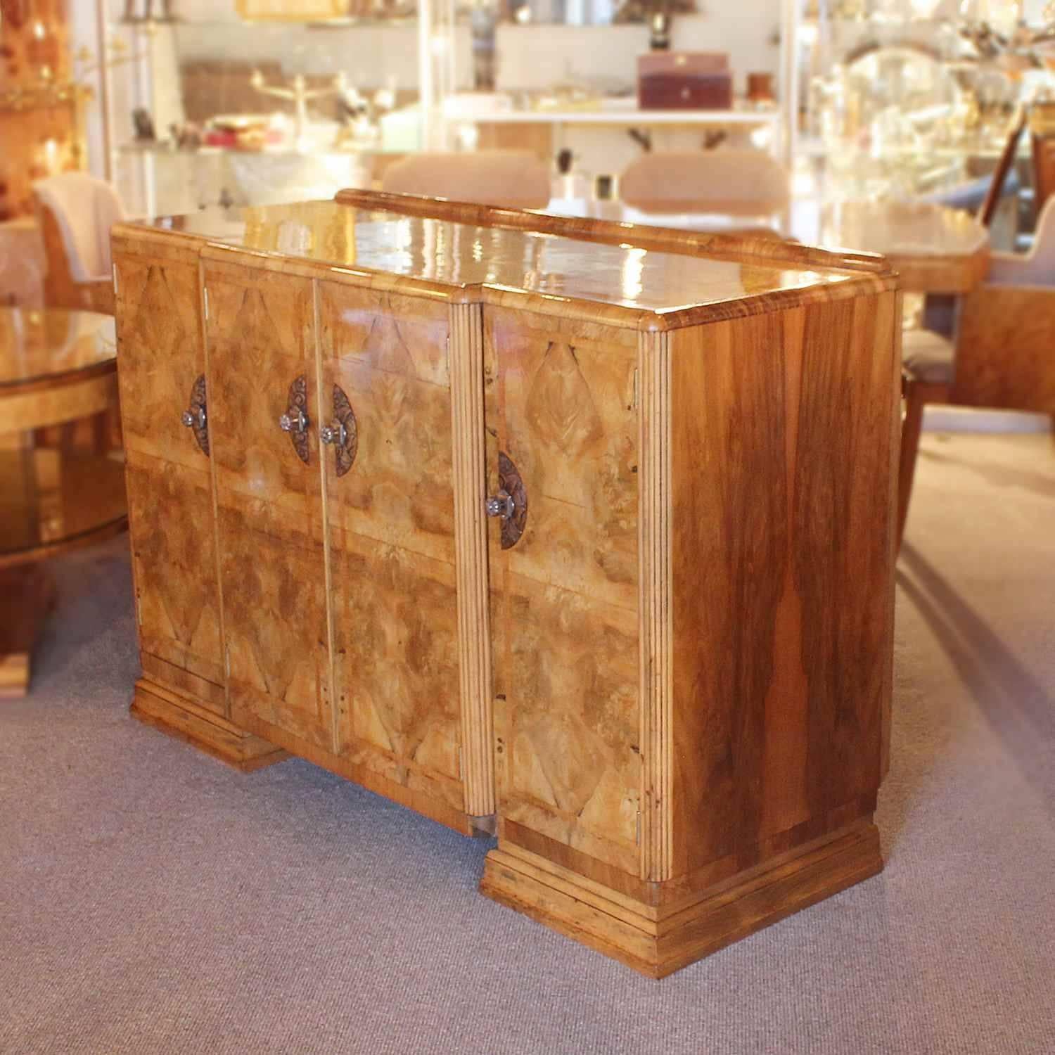 An Art Deco cocktail sideboard in walnut with satin birch banding, set over stepped bases. Central cabinet with two drawers and integral, rotating bottle holder. Carved oak and detailed brass handles.
 