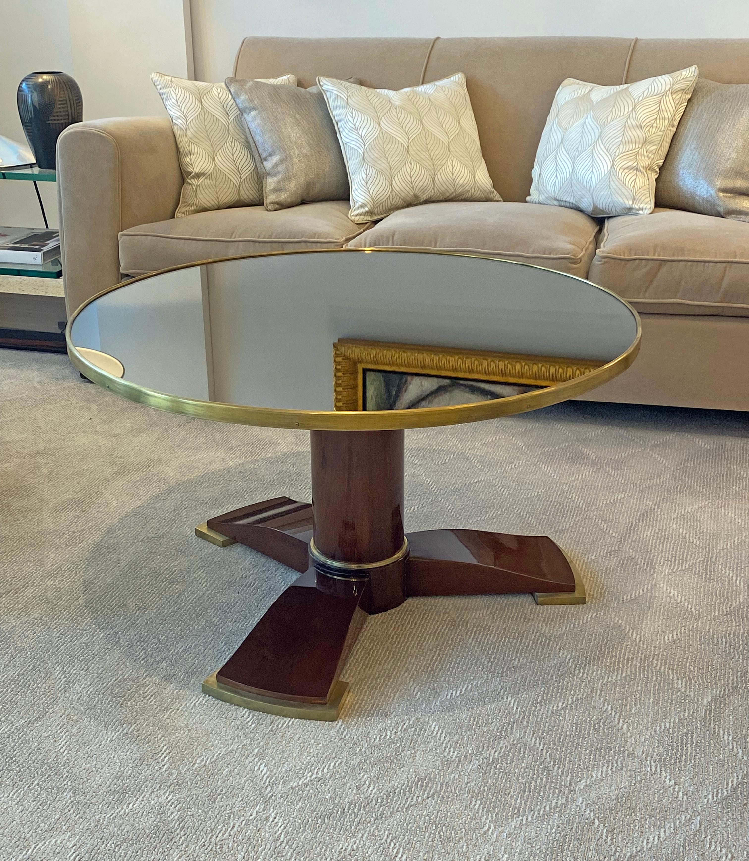 A round coffee table made from Rosewood with a circular top set with a mirrored glass panel raised on a cylindrical standard with outset base set with a Bronze band and continuing to three flipper-form feet. This table was made in France and