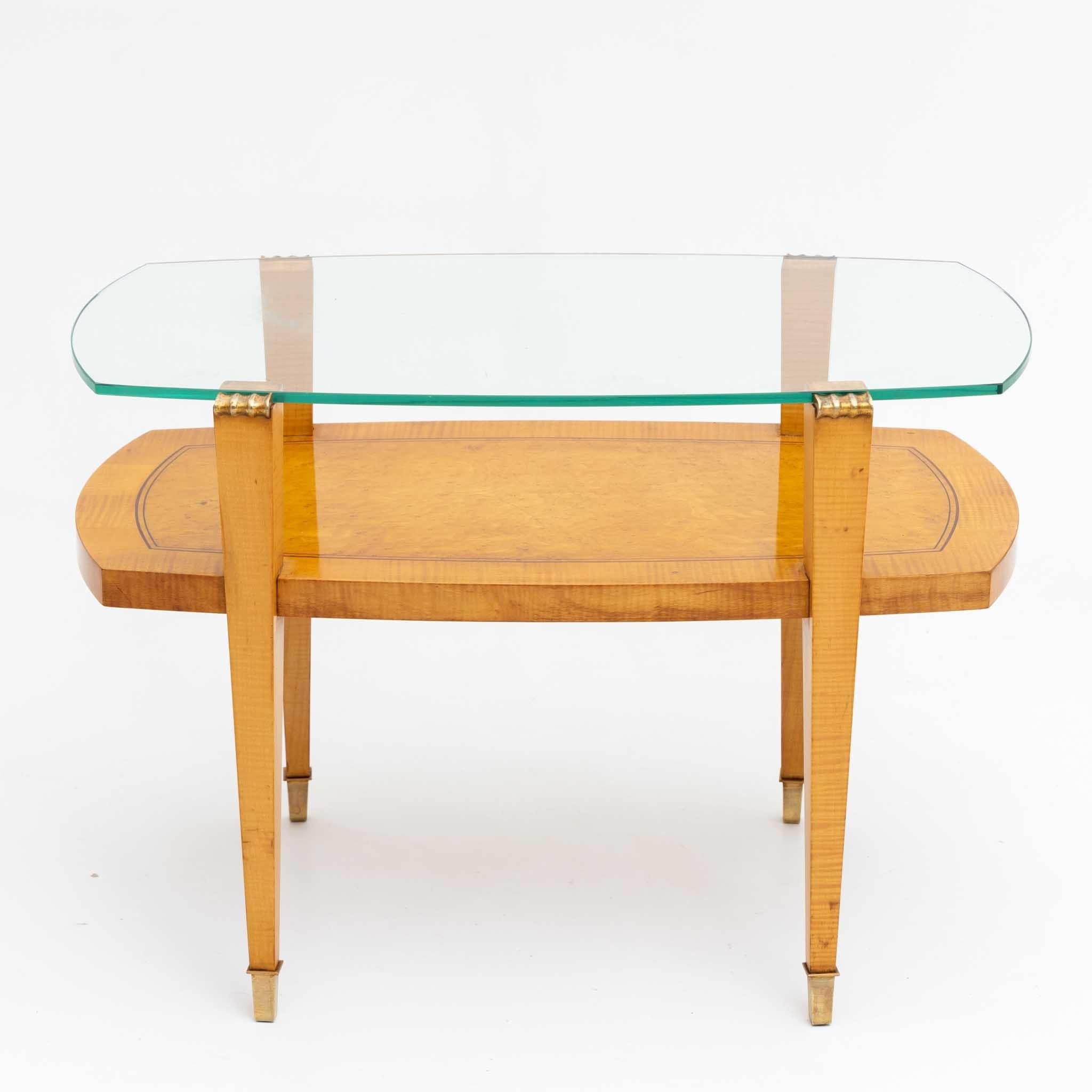 Coffee table with glass top on square tapering legs with brass sabots and intermediate shelf. The glass top is rounded on the narrow sides, as is the table top underneath. The table is veneered in Birch and hand polished.