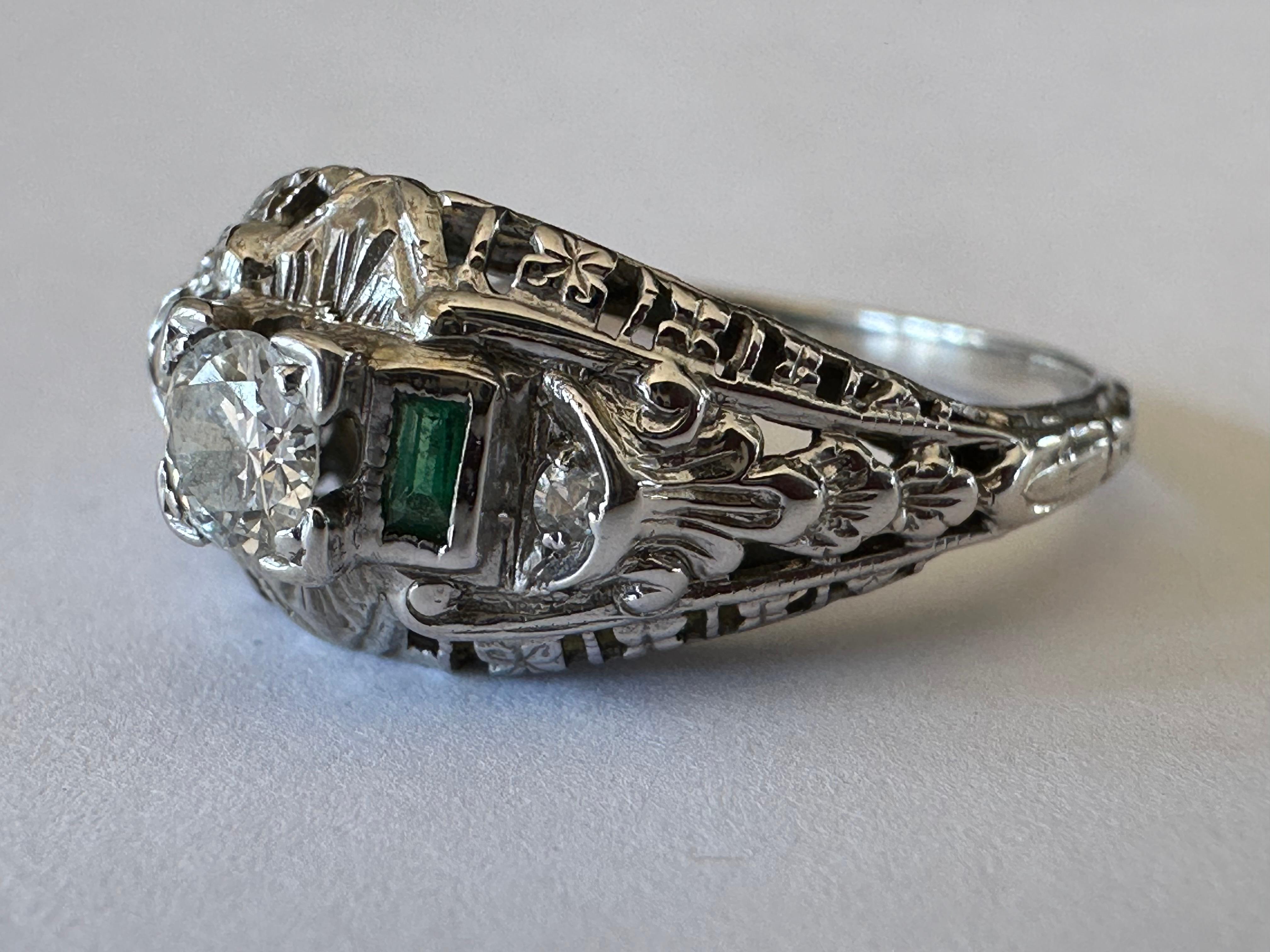 Crafted in 18K white gold, a fully-engraved shank and intricate piercing create a fanciful mounting for a shimmering Old European cut diamond center stone measuring approximately 0.20 carat, I color, VS clarity, complemented with two green Columbian