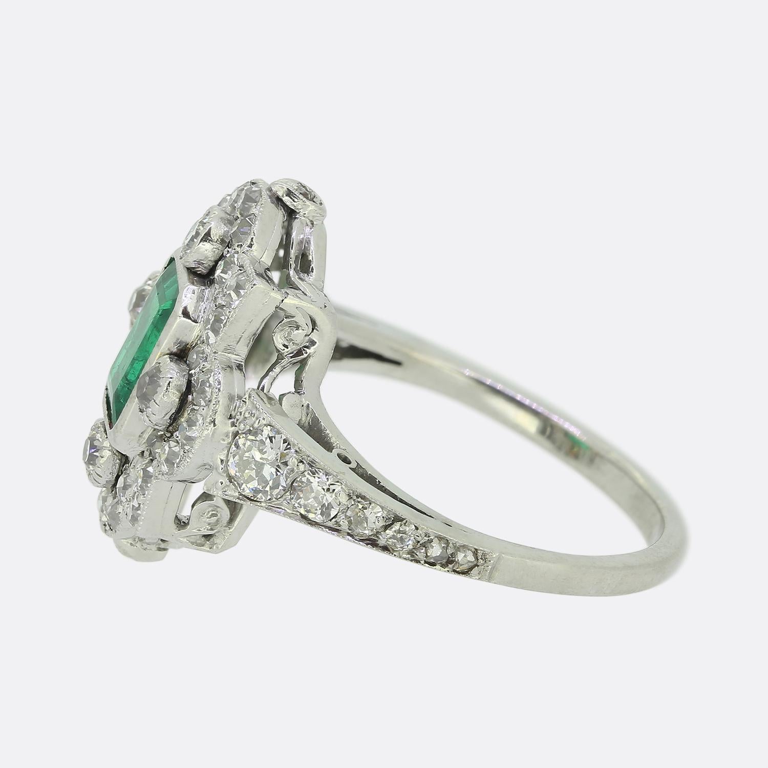 Here we have a marvellous emerald and diamond cluster ring crafted during the pinnacle of the Art Deco movement. This platinum piece features a single emerald cut emerald which sits slightly risen at the centre of the face. This principal stone is