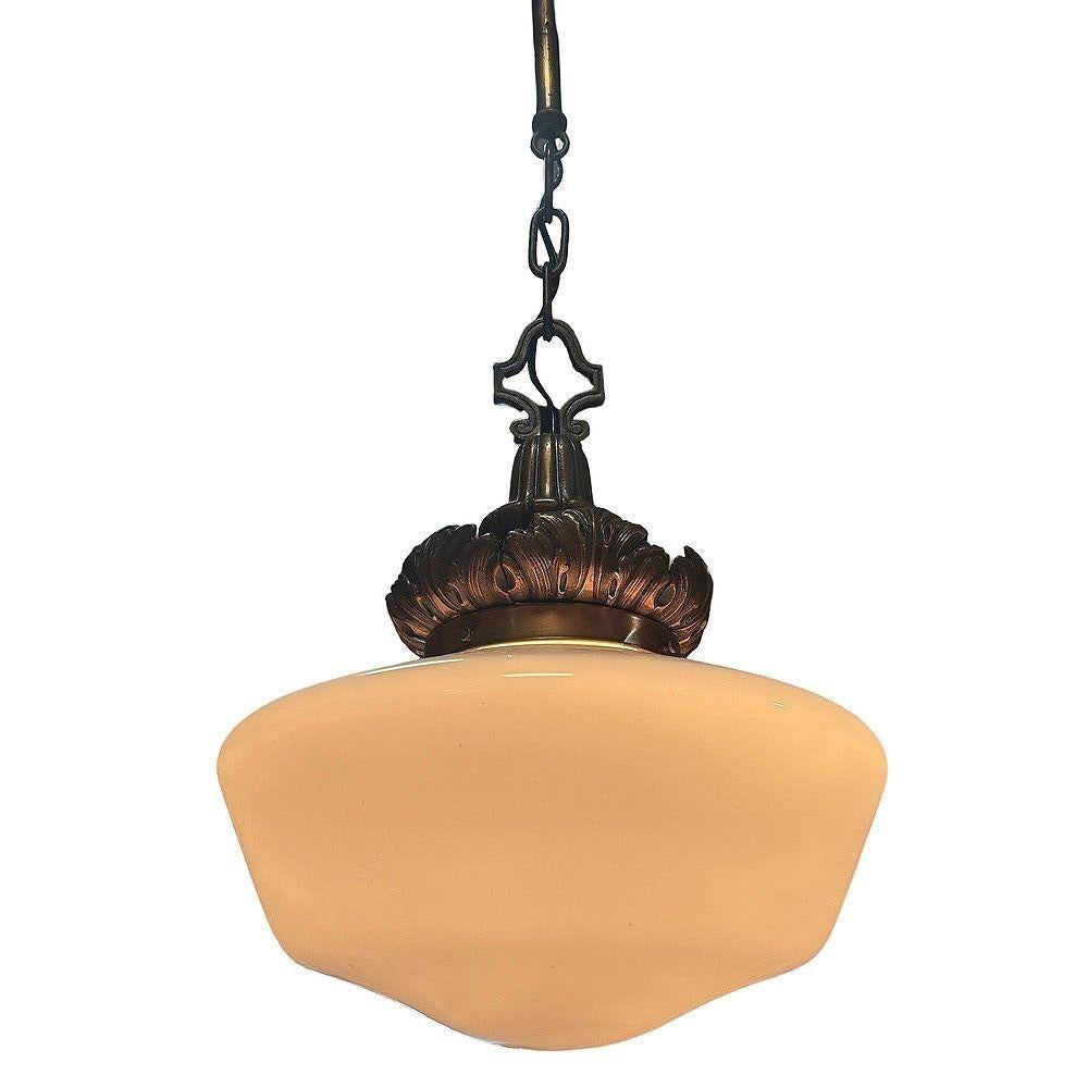 This 1930s Art Deco pendant light features a classic opal glass schoolhouse globe paired with an ornate cast brass fitter in a warm copper finish. The crown showcases acanthus leaf detailing, lending a refined neoclassical touch. A striking