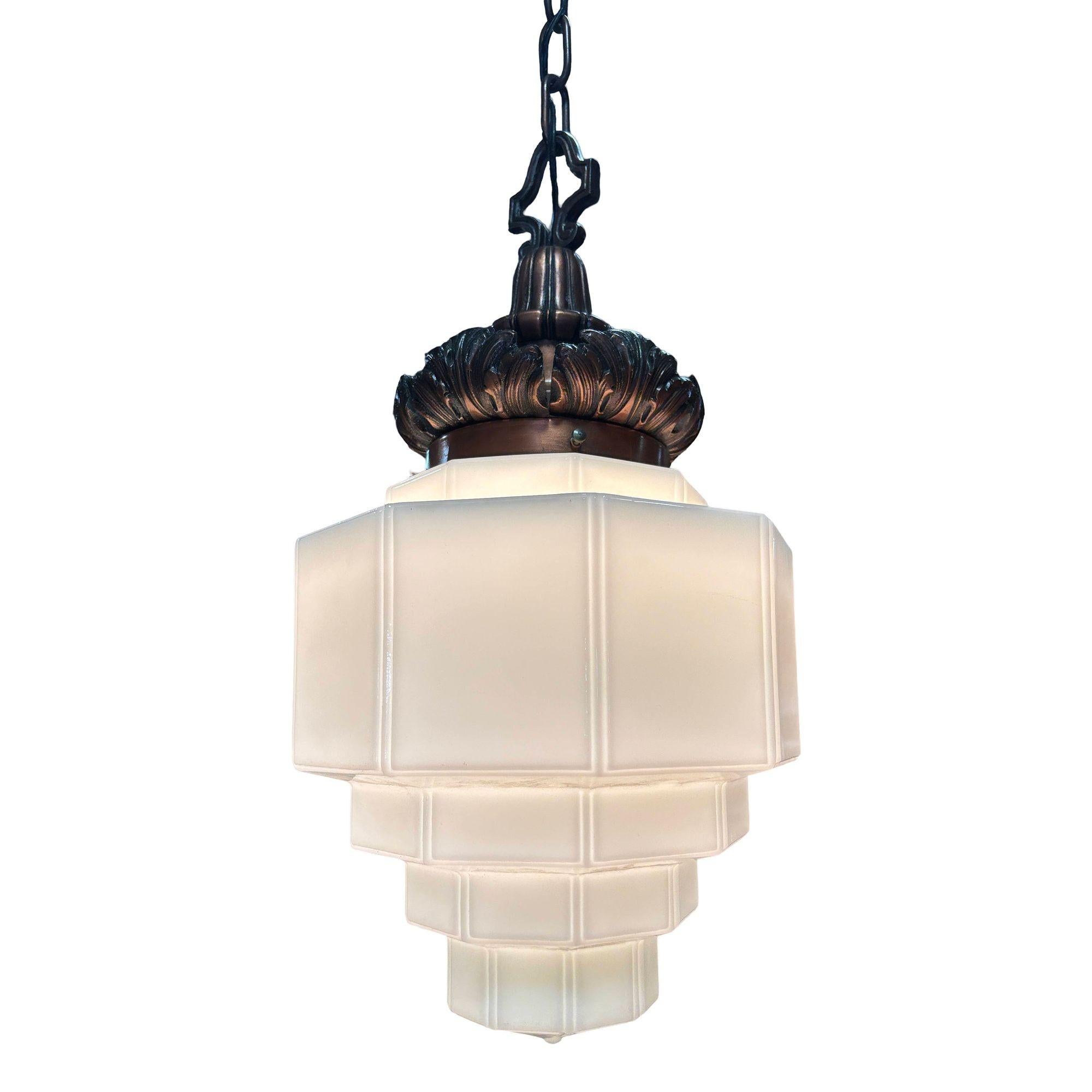 Lampada a sospensione Art Deco degli anni '30 con paralume in vetro lattiginoso a gradini e montatura in ottone fuso con finitura in rame caldo. La corona presenta dettagli a foglia d'acanto che aggiungono un tocco di eleganza neoclassica. Un