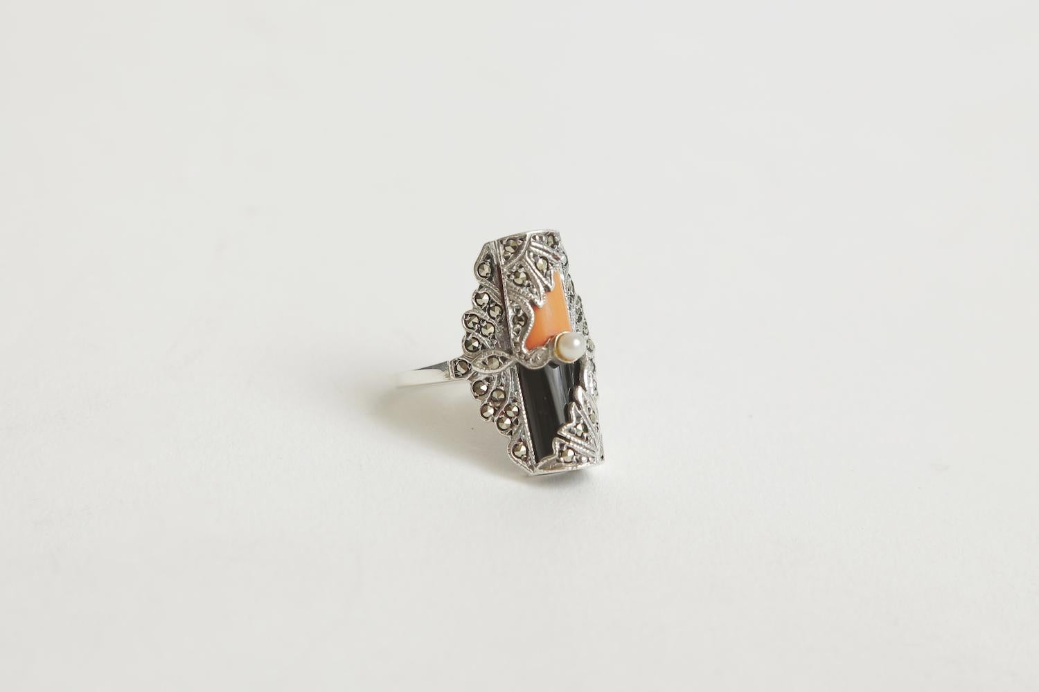 This lovely art deco ring is sterling silver and the design is set with surround marquisettes with angled coral and onyx and topped with a tiny tiny real pearl. It has a flair and is of the period. It is more elongated and slender on the finger.