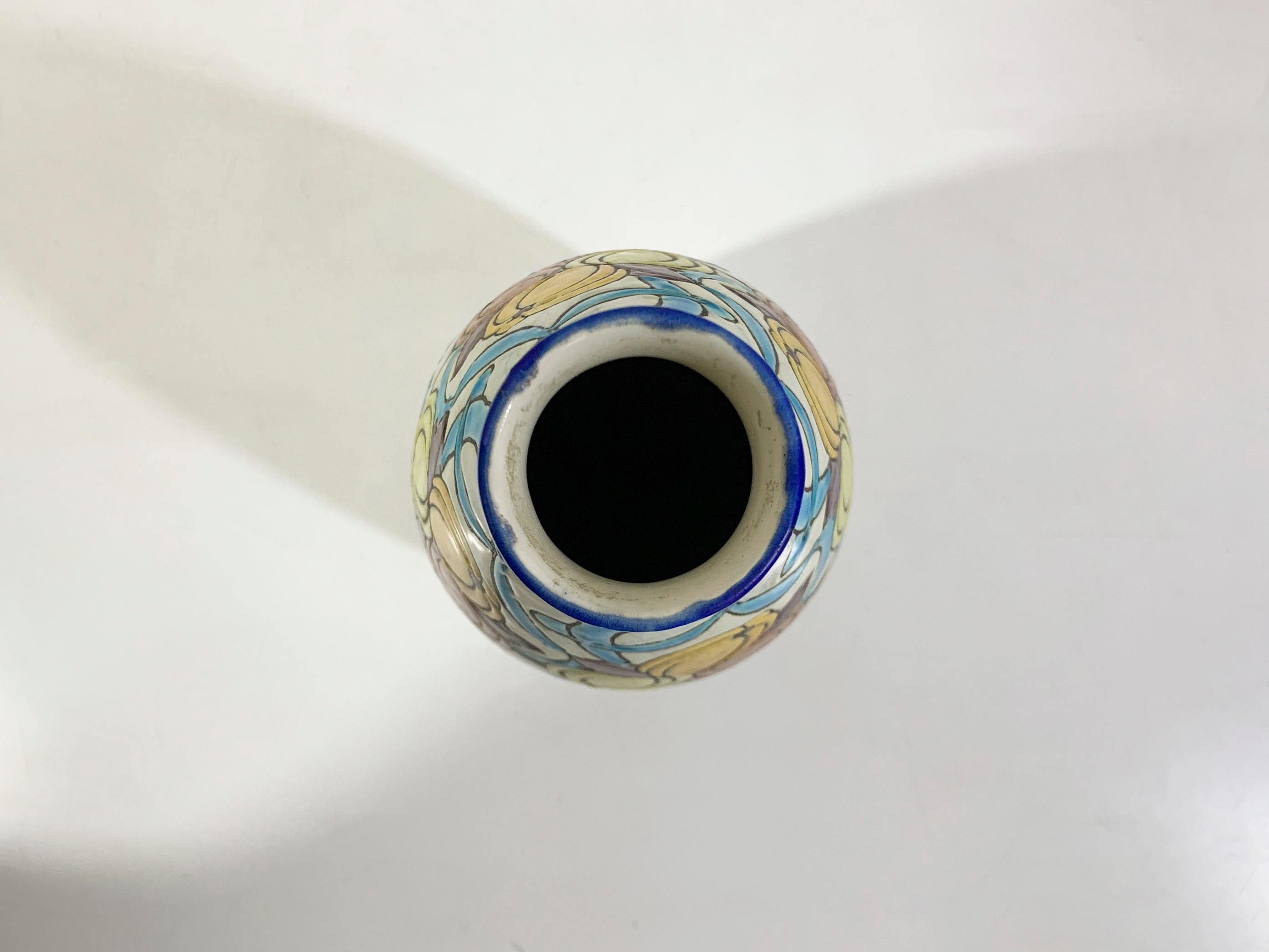 Ce vase estampillé Boch La Louvière présente un magnifique motif floral peint à la main avec des fruits jaunes, orange et rouges vibrants entrelacés avec des motifs de rubans bleus, sur un fond de glaçure craquelée blanc cassé. Sa forme arrondie et