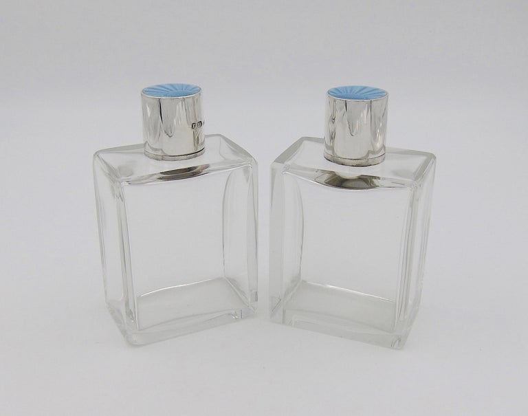 A pair of English Art Deco perfume bottles with sterling silver and guilloche enamel tops by Finnigans, Ltd., date marked 1936. The rectangular scent bottles have beveled edges and both retain their original glass stoppers. The sterling silver