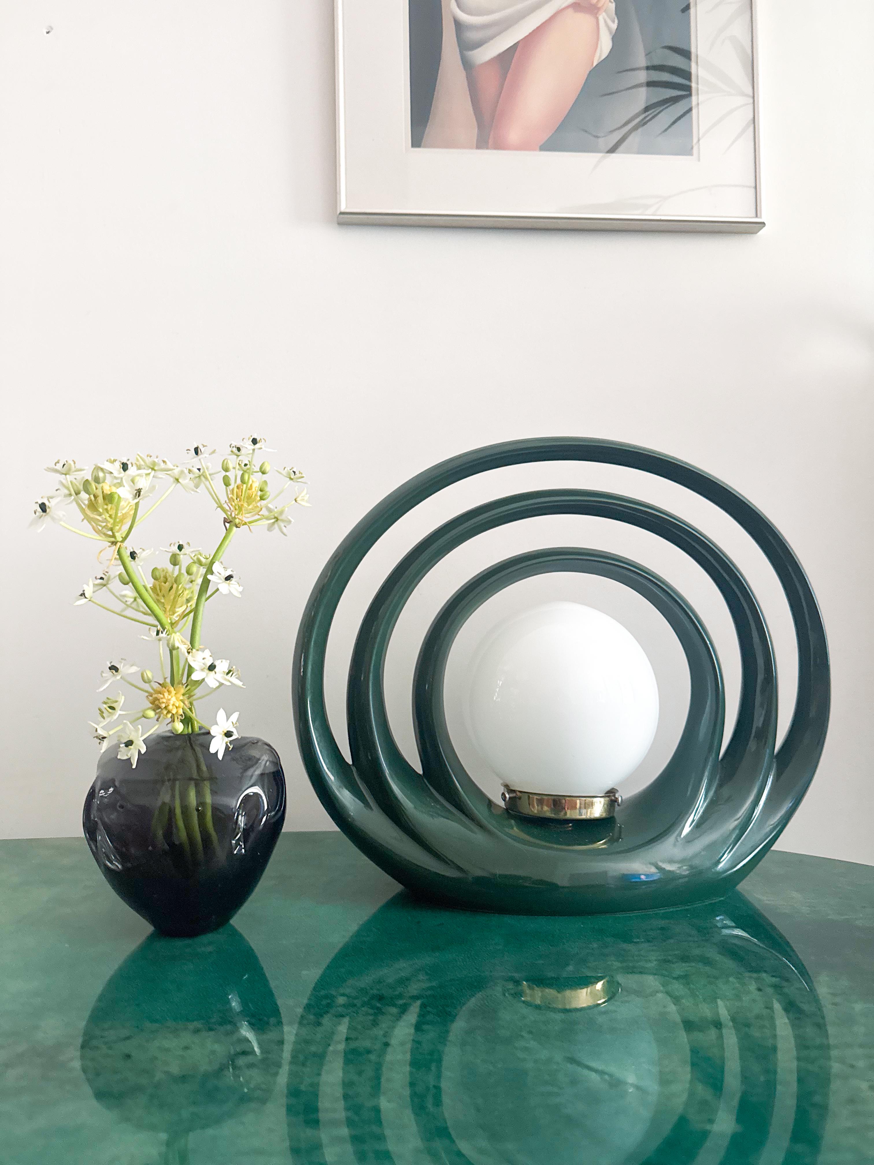 An extraordinary Art Deco–inspired halo lamp, crafted in richly glazed dark green ceramic with a sculptural circular form. The bold, architectural design features a series of concentric arcs framing a central white glass globe, elevated by a