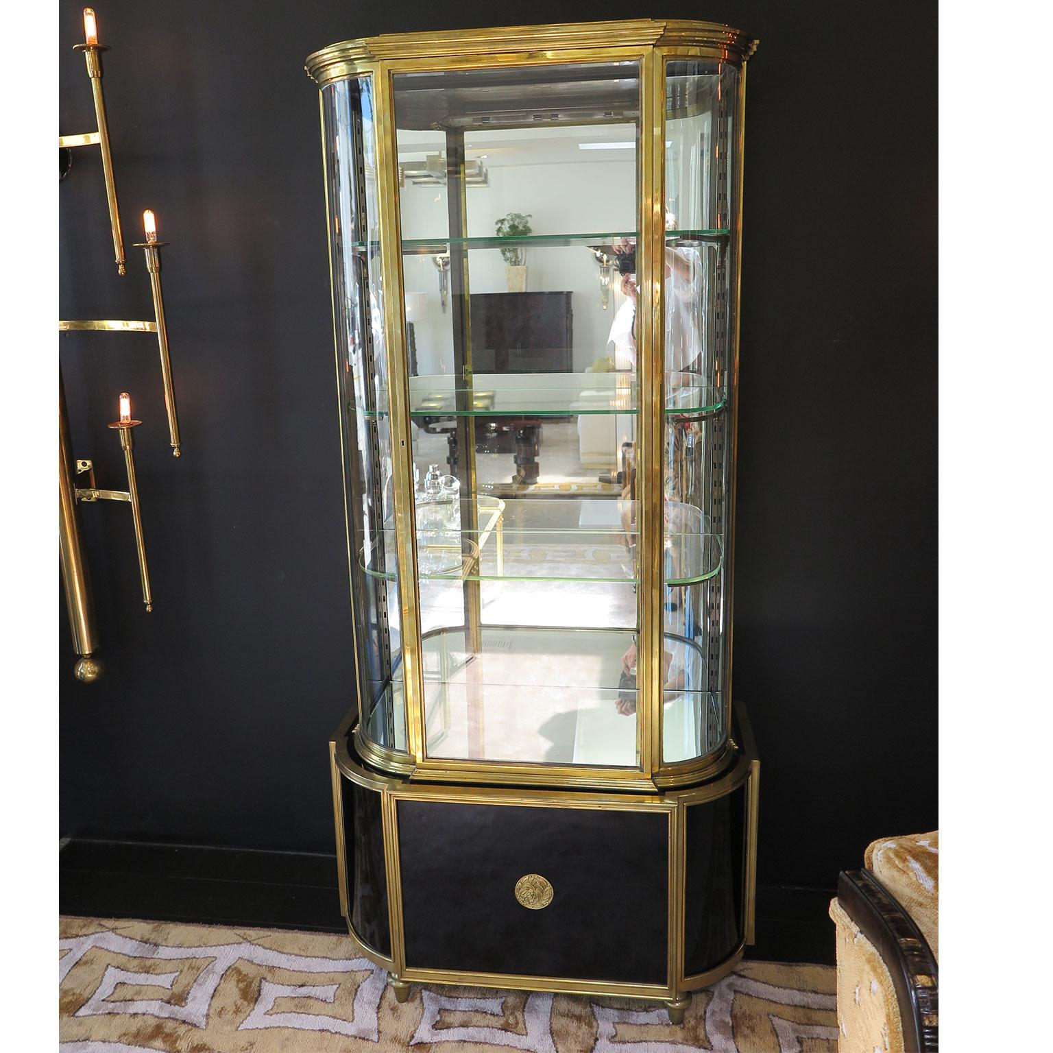 This tall and stately Art Deco brass display cabinet features a mirrored interior back and curved glass. Three adjustable interior glass shelves offer ample storage space for displaying collections. A concealed light bar illuminates the interior of