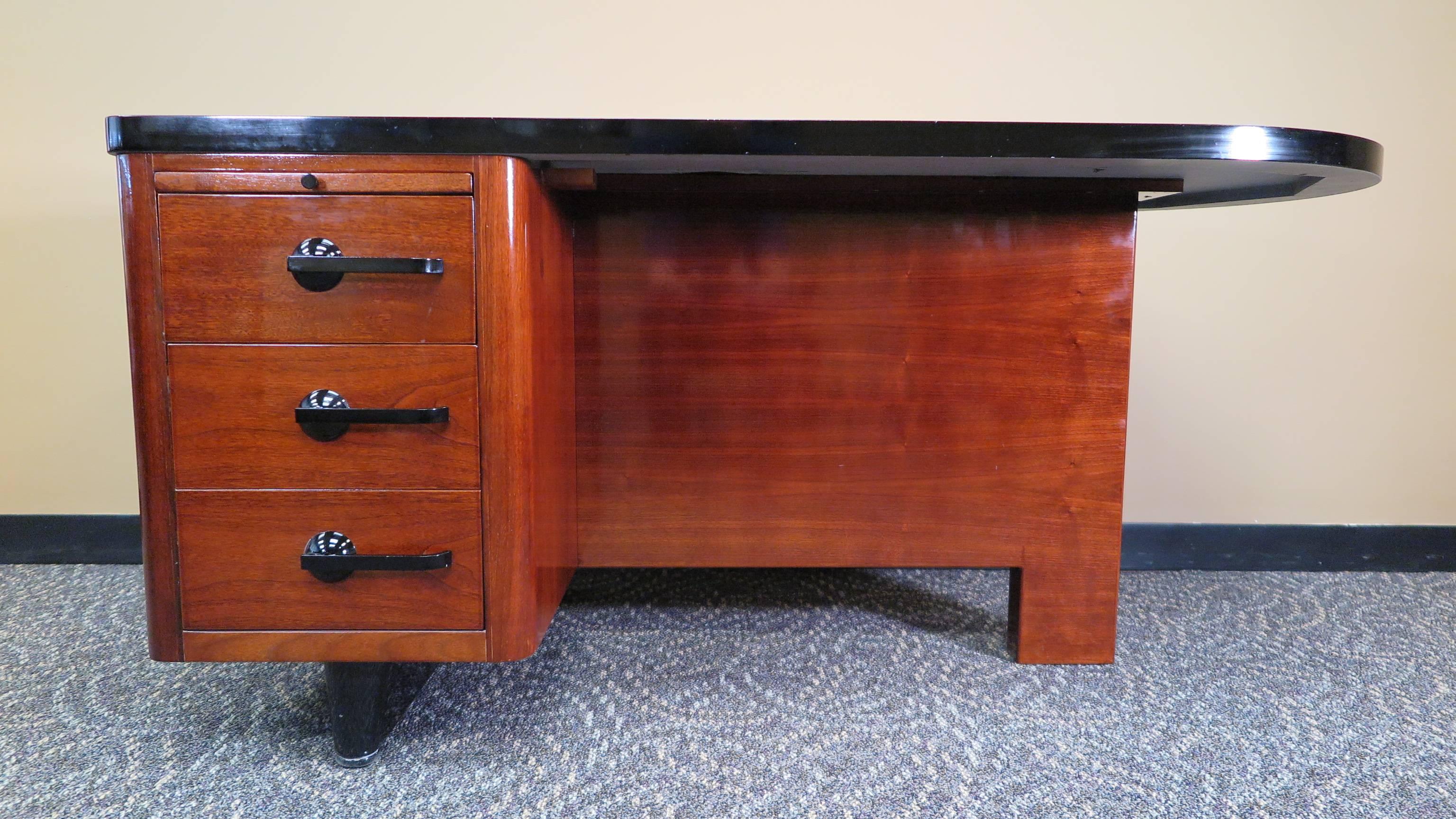 An Art Deco desk in the style of Gilbert Rhode. Mahogany Art Deco desk with arched shape top to one side. Writing platform on left side with one drawer and a file deep drawer lower.  In very good condition chair pictured is included.
