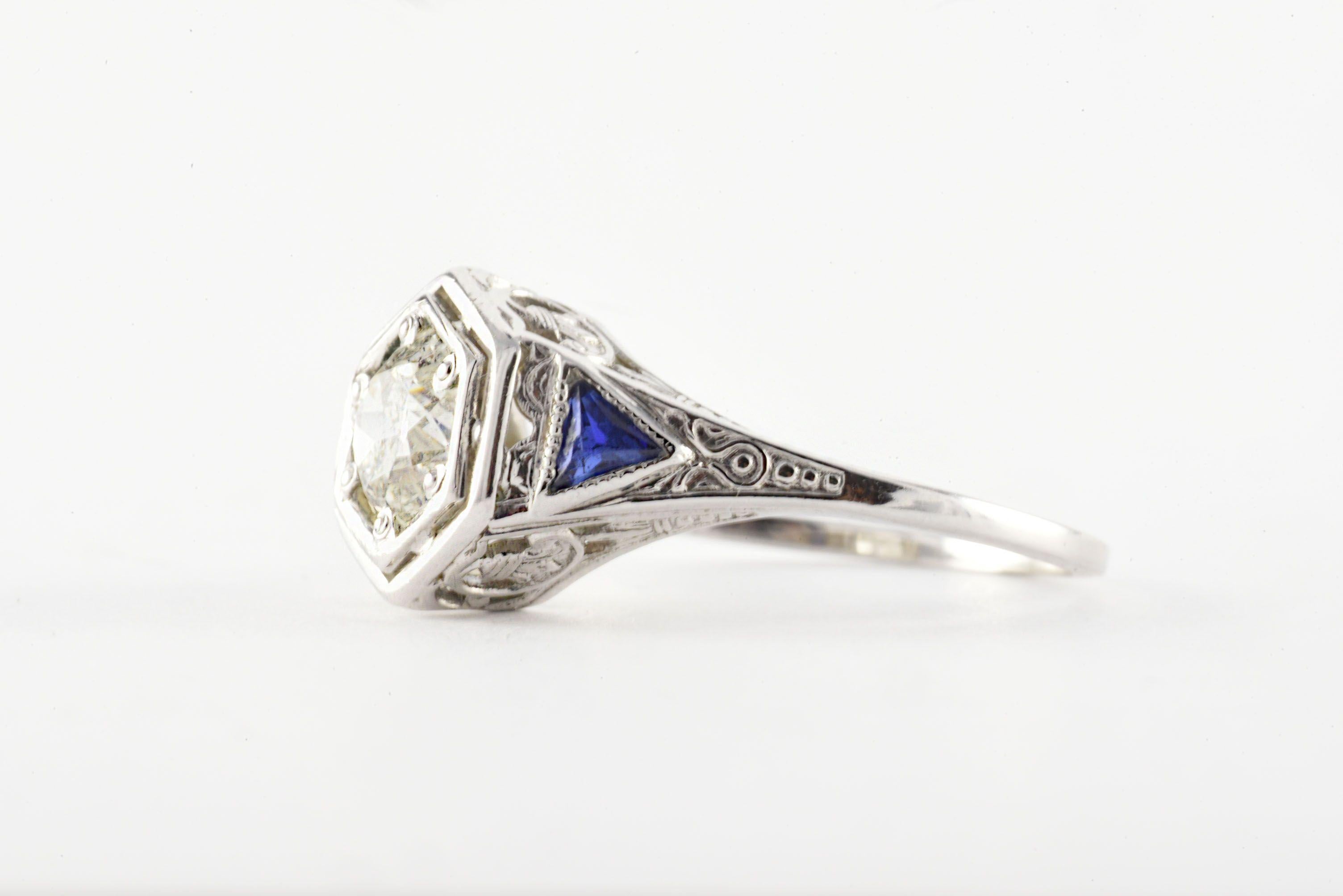 Crafted in the 1920-30s from 18kt white gold, this Art Deco gem features a bold Old European cut diamond measuring approximately 0.75 carats, H-I color, I1 clarity, and complemented with two matching trillion blue sapphires and intricate filigree. 
