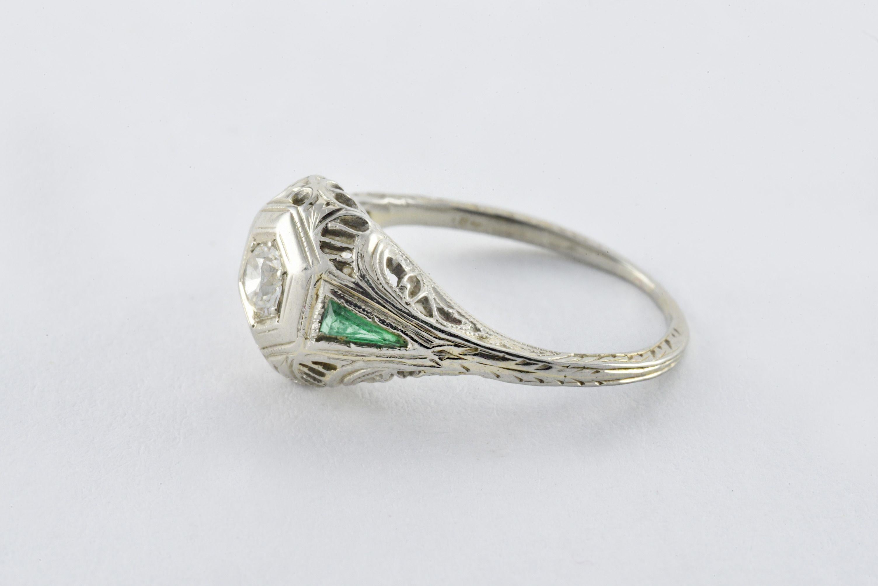 An Old European cut diamond measuring approximately 0.25 carats sits at the center of this classic Art Deco gem accented with two trillion cut green emeralds and fine filigree and mounted in 18kt white gold. 
