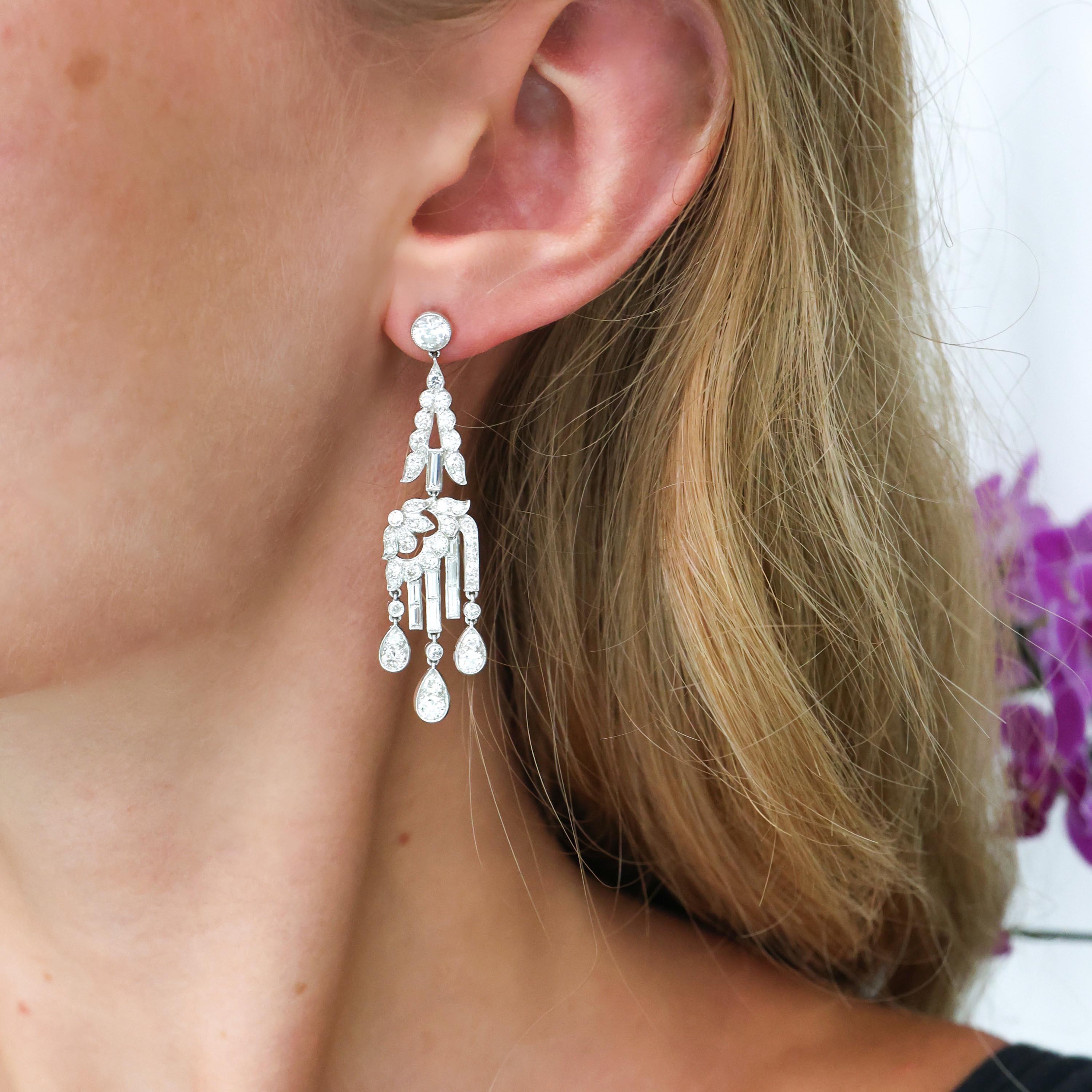 A pair of Art Deco diamond and platinum drop earrings, with rub over set round brilliant-cut diamond tops, adjoining pendants with a scalloped inverted V shape, holding a baguette-cut diamond, linked to a  in a stylised, articulating design of a
