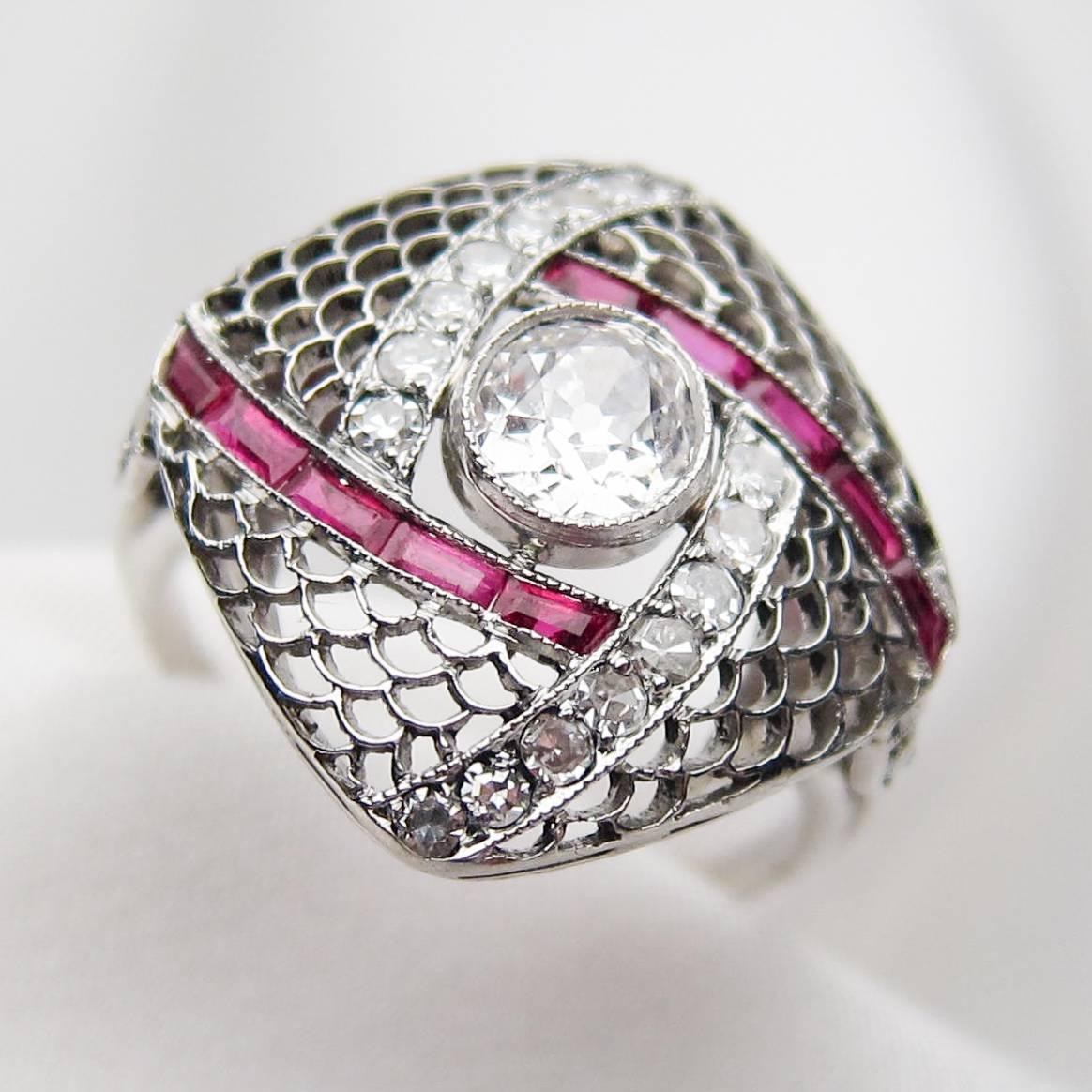 Circa 1930. This intriguing platinum Art Deco filigree ring features the dynamic contrast between icy diamonds and lush rubies. The diagonal, square cushion-shaped top of the ring features a central, bezel-set, old European-cut diamond weighing .53