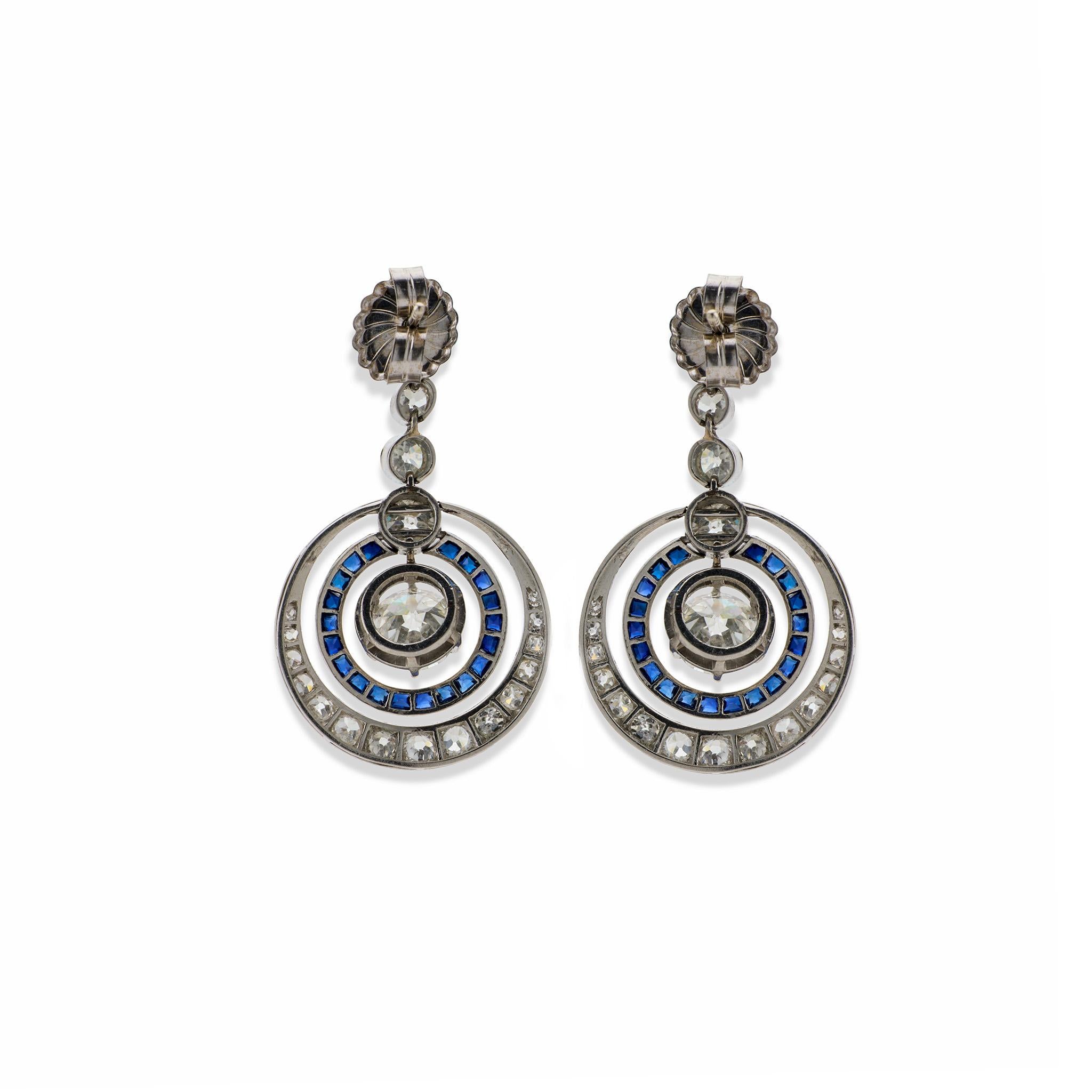 Dating from the 1920s. these Art Deco pendant earrings are composed of platinum, sapphires and diamonds. Each pendant earring is designed as a cascade of round-cut diamonds, the lowest encircled by freely moving, concentric hoops of diamonds and