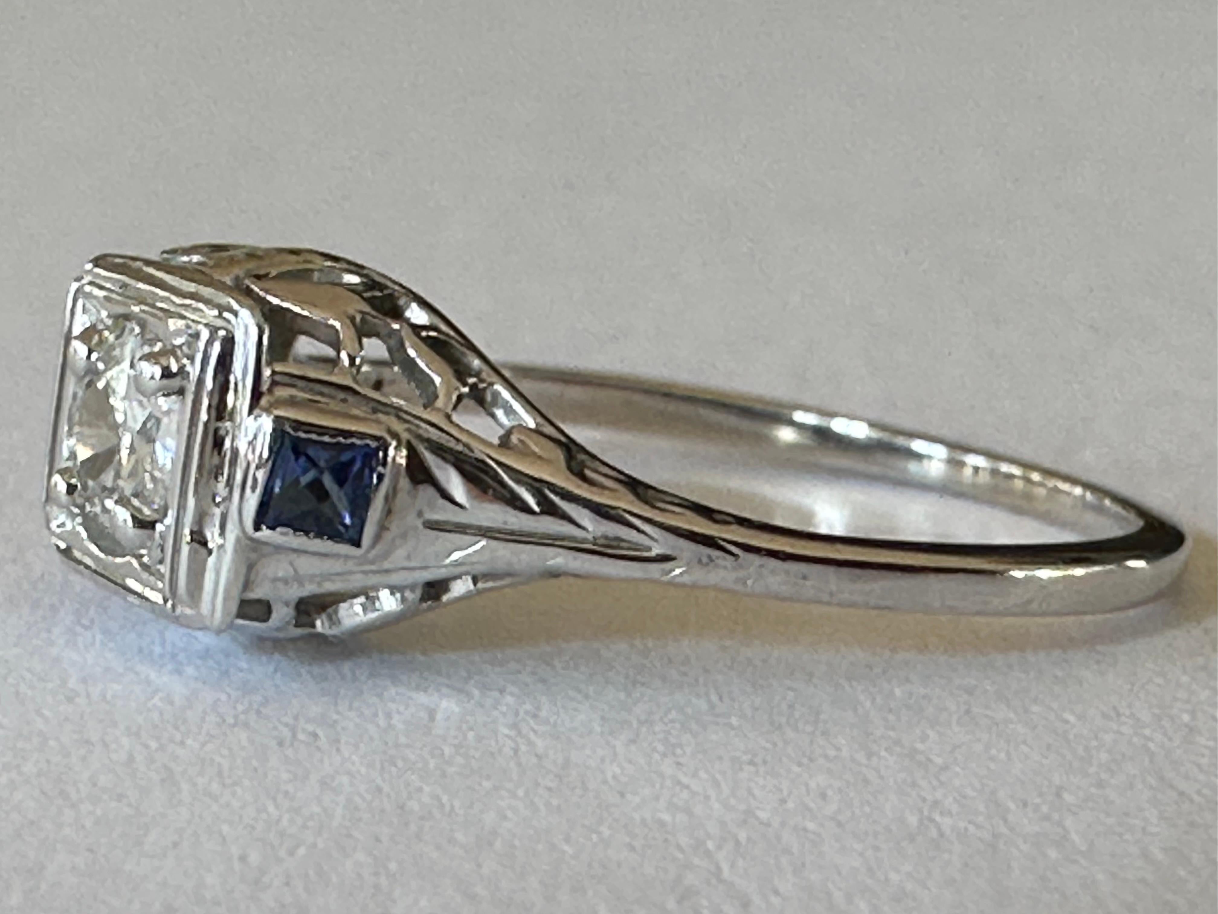 A bold 0.25-carat Old European cut diamond center stone, GH color, SI2 clarity is complemented with two  blue sapphires and intricate filigree in this stunning Art Deco ring handcrafted in 18K white gold. 