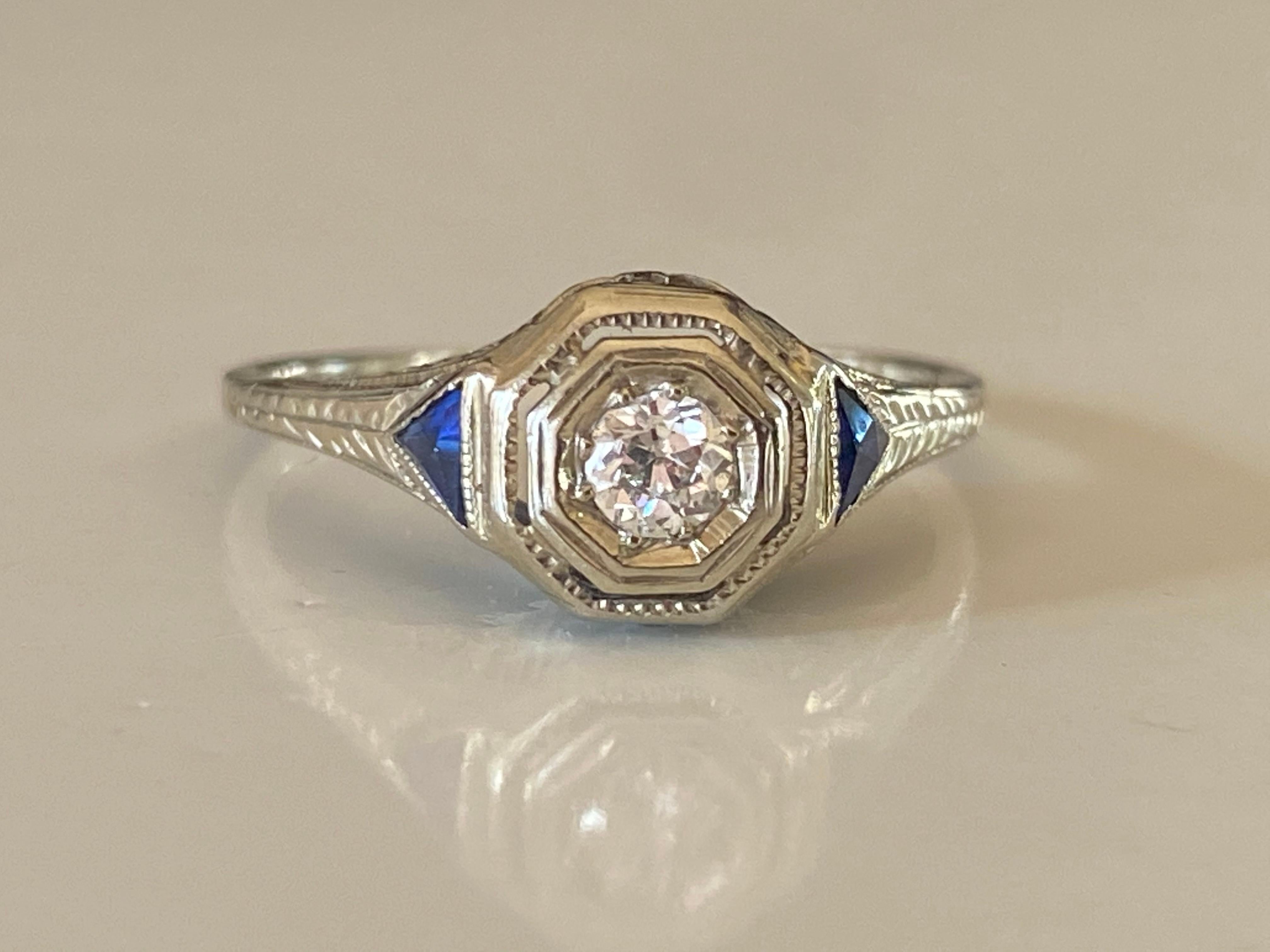 Crafted in the 1920s in 18kt white gold, this classic Art Deco gem is designed around a sparkling Old European cut center stone measuring approximately 0.15 carts, G color, SI1 clarity and complemented with two blue triangle sapphires and intricate