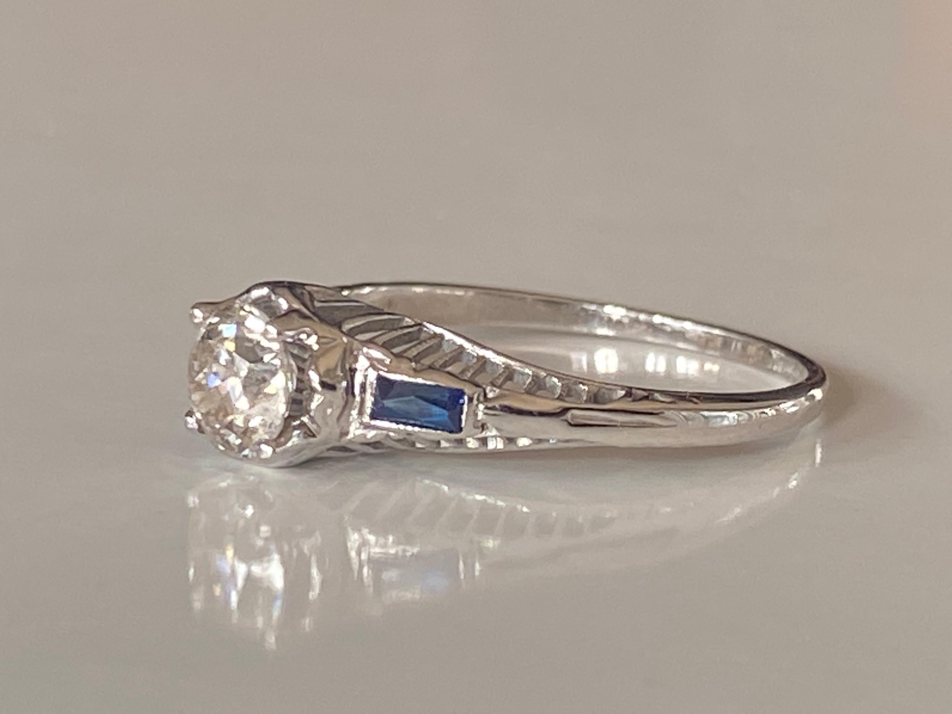 This antique Art Deco ring crafted in the 1920s features an Old European cut diamond center stone measuring approximately 0.65 carats, J color, SI1 clarity, complemented by two baguette blue sapphire side stones set in an 18kt white gold filigree
