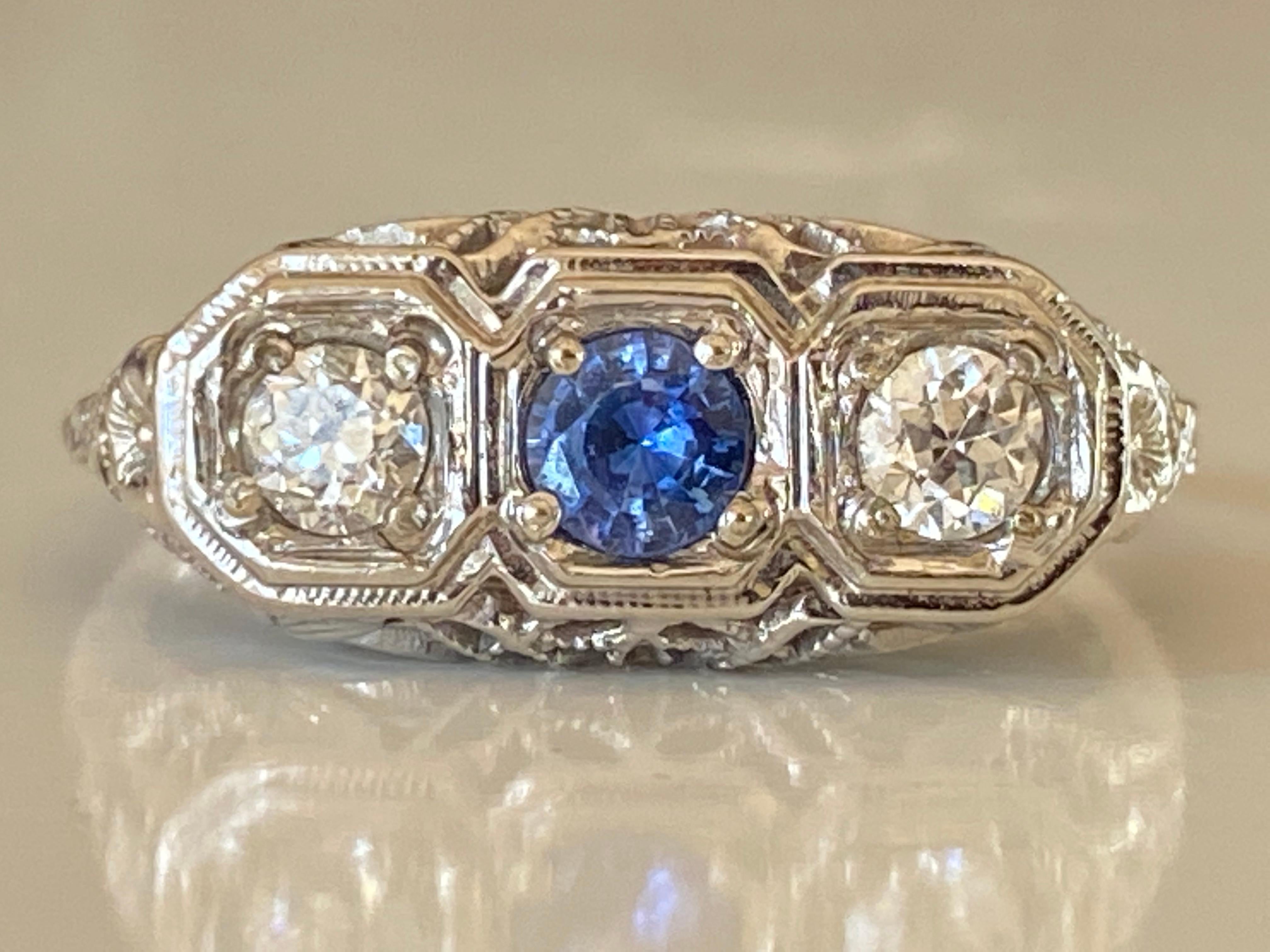 This antique Art Deco three-stone ring handcrafted in 18kt white gold features a stunning blue sapphire center stone flanked by two Old European cut diamonds totaling 0.30 carats, H color, SI clarity accented with fine filigree details and floral