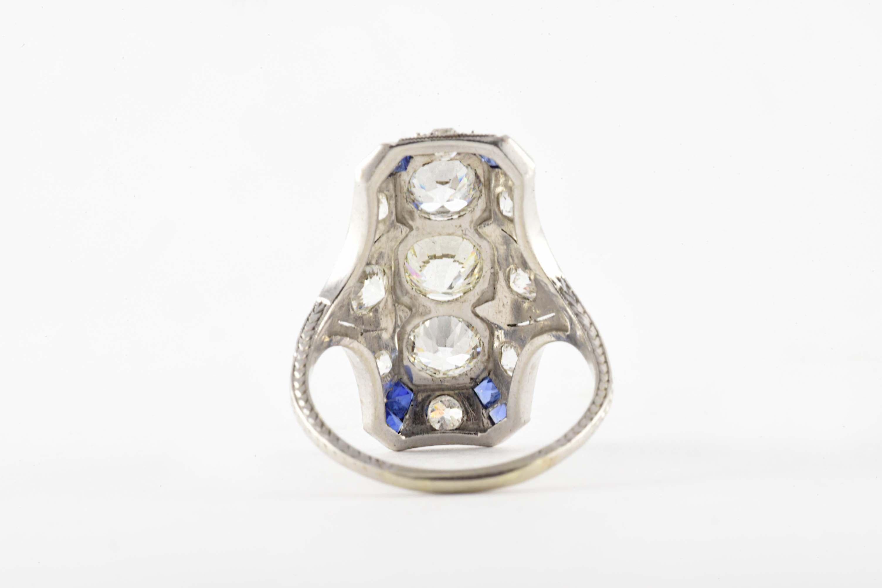 This glistening 14kt white gold and platinum ring is designed around a trio of Old European cut diamonds each measuring 0.65 carats, F-G color, VS clarity, accented with eight smaller Old European cut diamonds and eight calibrated blue square-shaped