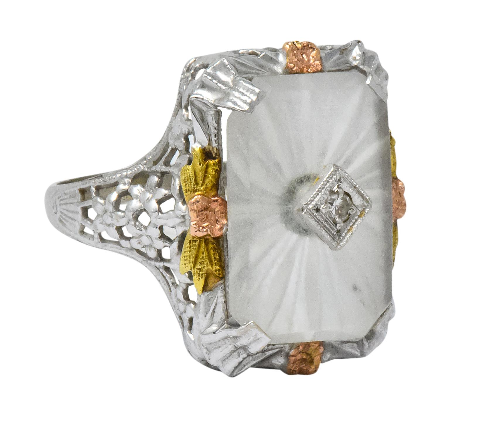Centering a rectangular camphor glass tablet with a radiating frosted design, inset with a single cut diamond

Prong set and surrounded by rose gold flowers with green gold leaves

White gold gallery with pierced floral and foliate design

Stamped