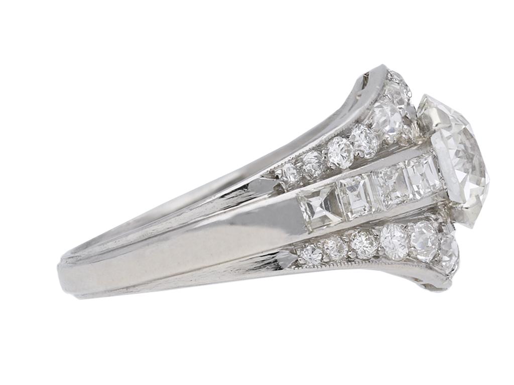 Art Deco diamond cluster ring. Set to centre with a round old cut diamond, H colour, VS2 clarity, with an approximate weight of 2.01 carats in an open back claw setting, flanked by eight square step cut diamonds in open back rubover settings with a