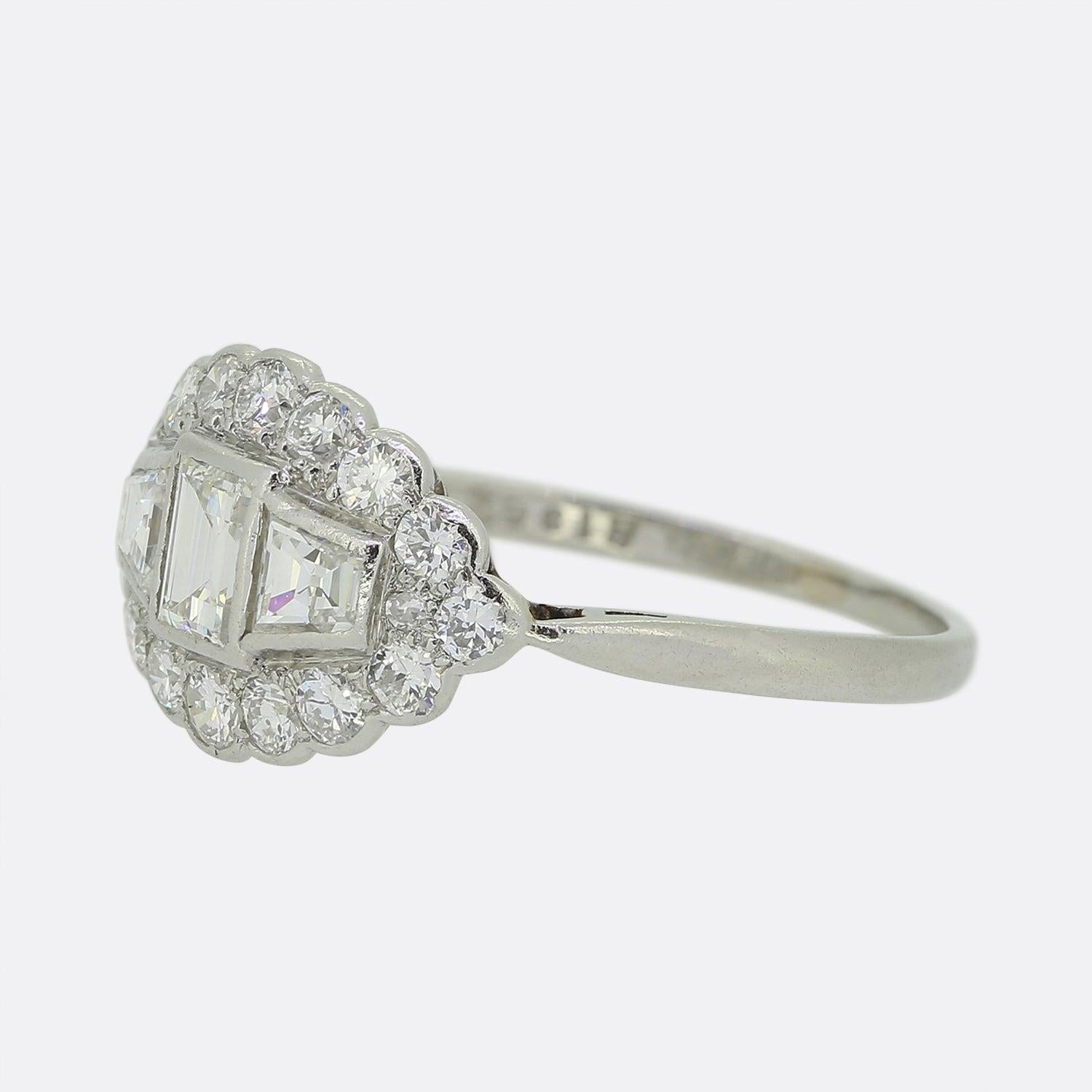 Ecco un meraviglioso anello con diamanti a grappolo risalente a un'epoca in cui lo stile Art Déco era l'approccio pionieristico al design. Un singolo diamante taglio smeraldo è incastonato al centro del quadrante e un singolo diamante taglio calibro