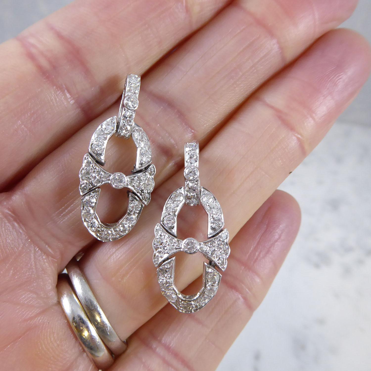 A pair of diamond drop earrings from the Art Deco period comprising a bar of old cut diamonds suspending an articulated, open oval loop with a central horizontal bar and single cut diamonds.  White claw settings.  Total estimated diamond content