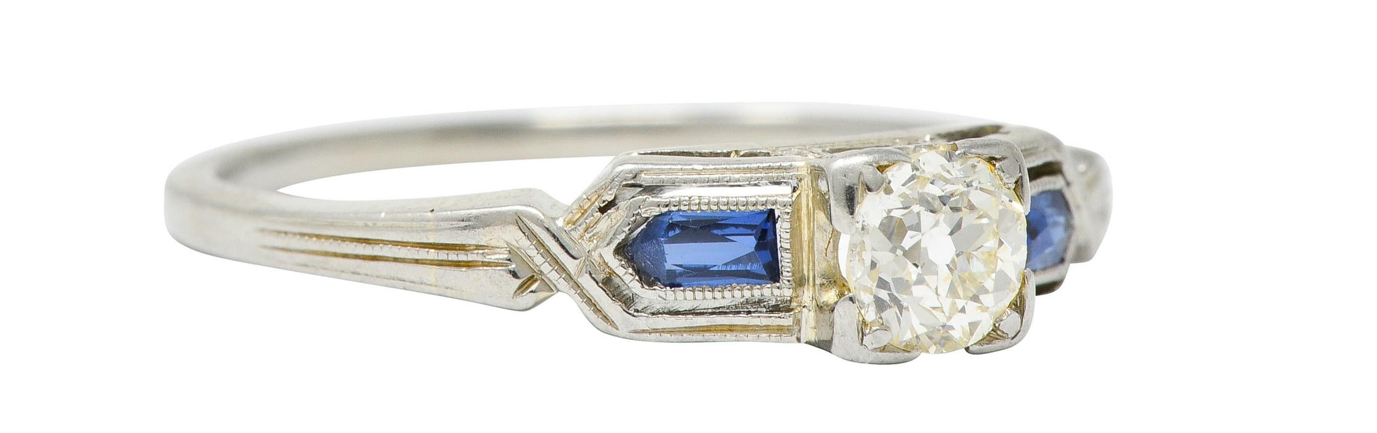 Centering an old European cut diamond weighing approximately 0.27 carat; K/L color and VS clarity

Set in an engraved square form head flanked by slight cathedral shoulders

With bright blue calibrè cut sapphires weighing in total approximately 0.10