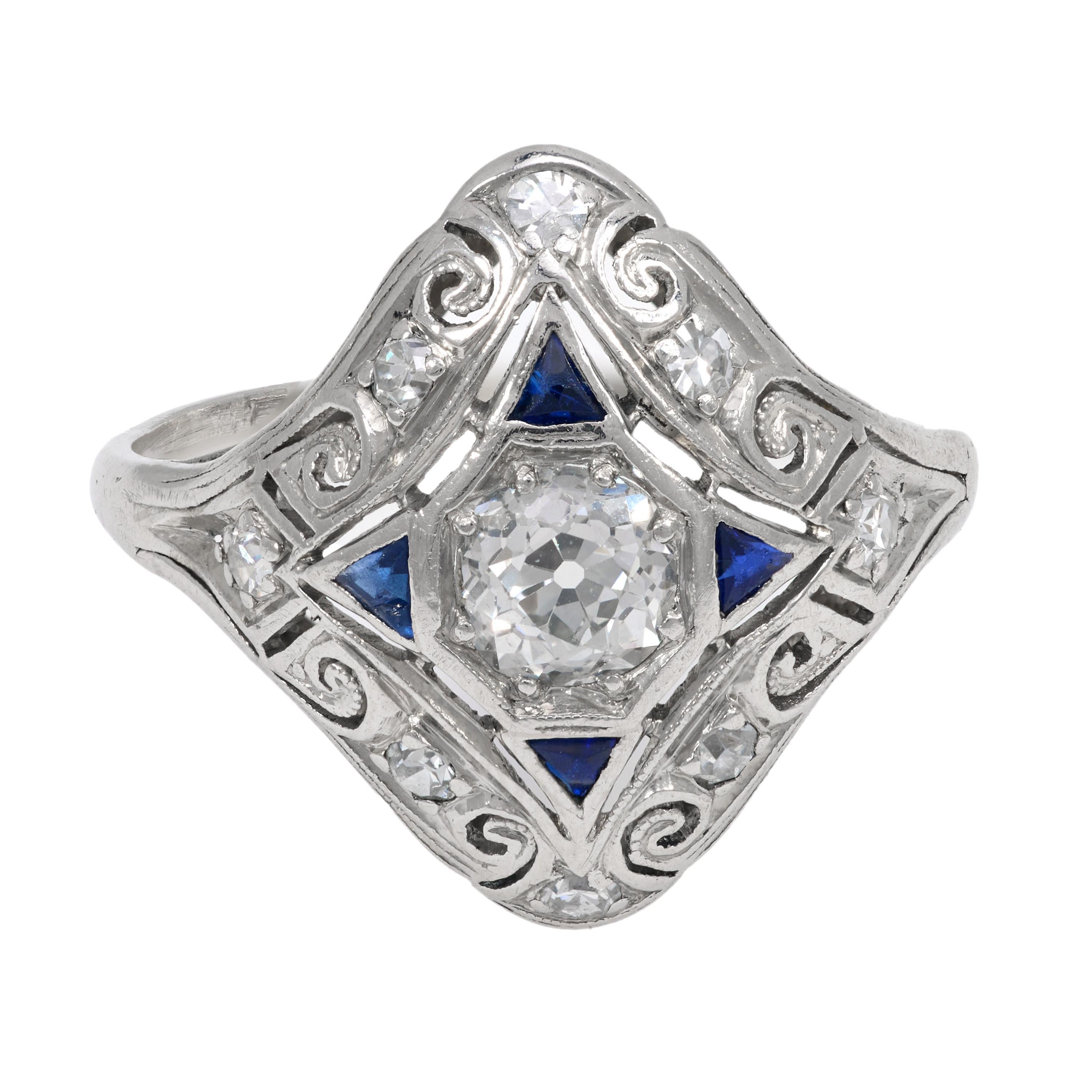 This platinum Art Deco shield ring dazzles with a .60 carat old European cut diamond framed by vibrant calibre cut sapphires and crisp single cut diamonds. Intricate filigree work and geometric elegance hallmark the 1930s era, making this